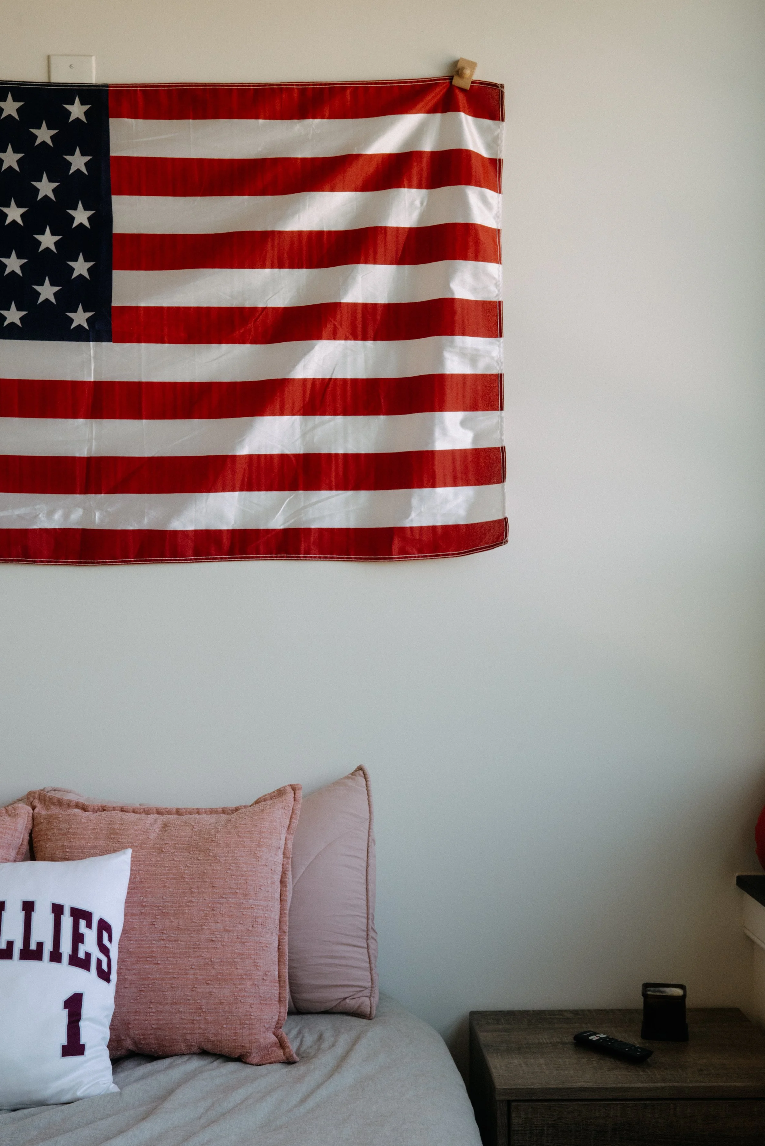 A wall decorated with an American flag hanging on a wall. There is a bed with pink and white pillows, one with the word 'BELLIES' and the number '1'. A nightstand with a remote control and a black speaker is next to the bed.