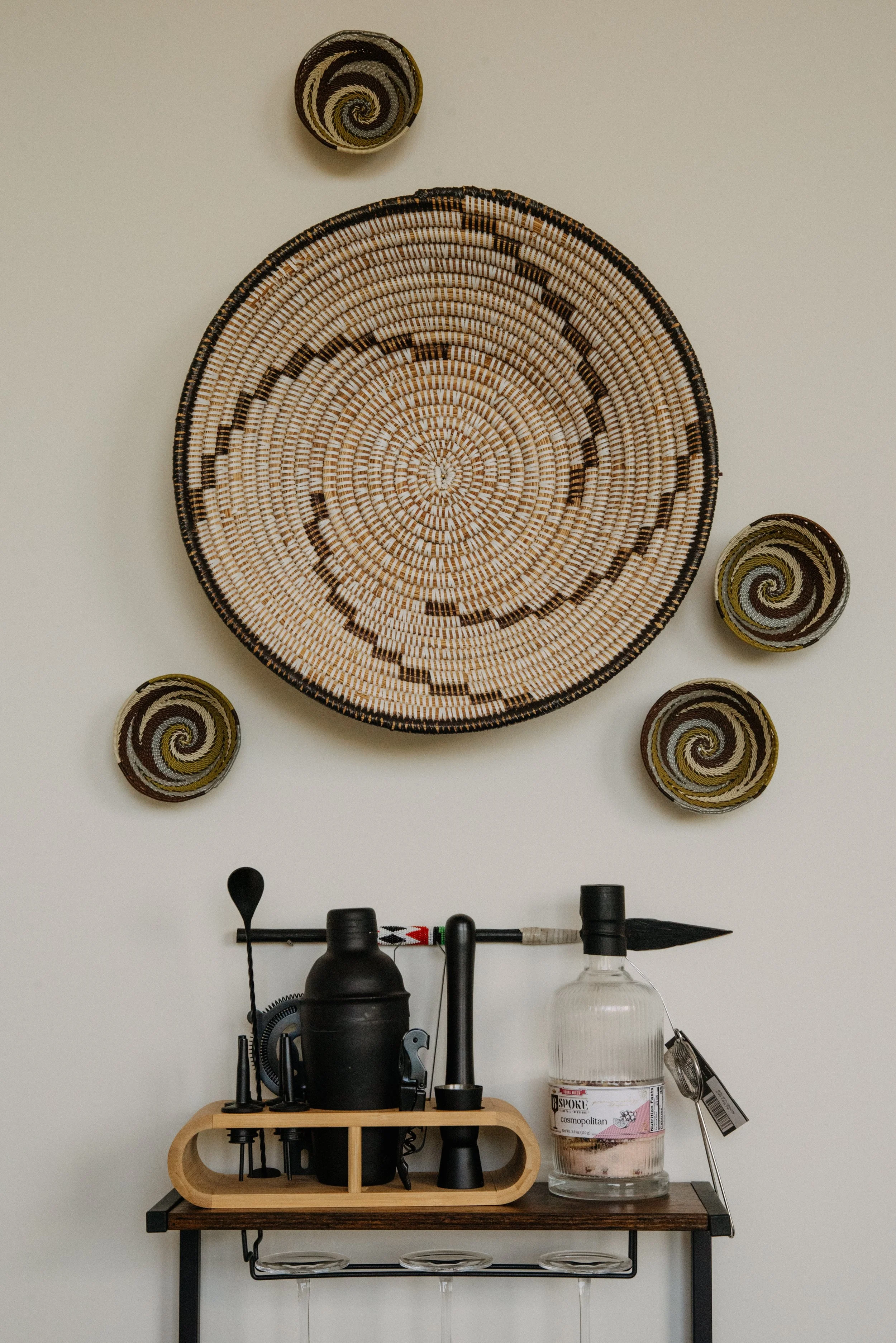Wall decorated with woven baskets, a wooden shelf holding bar tools, a large glass bottle, and a black container, with the wall color being a neutral off-white.