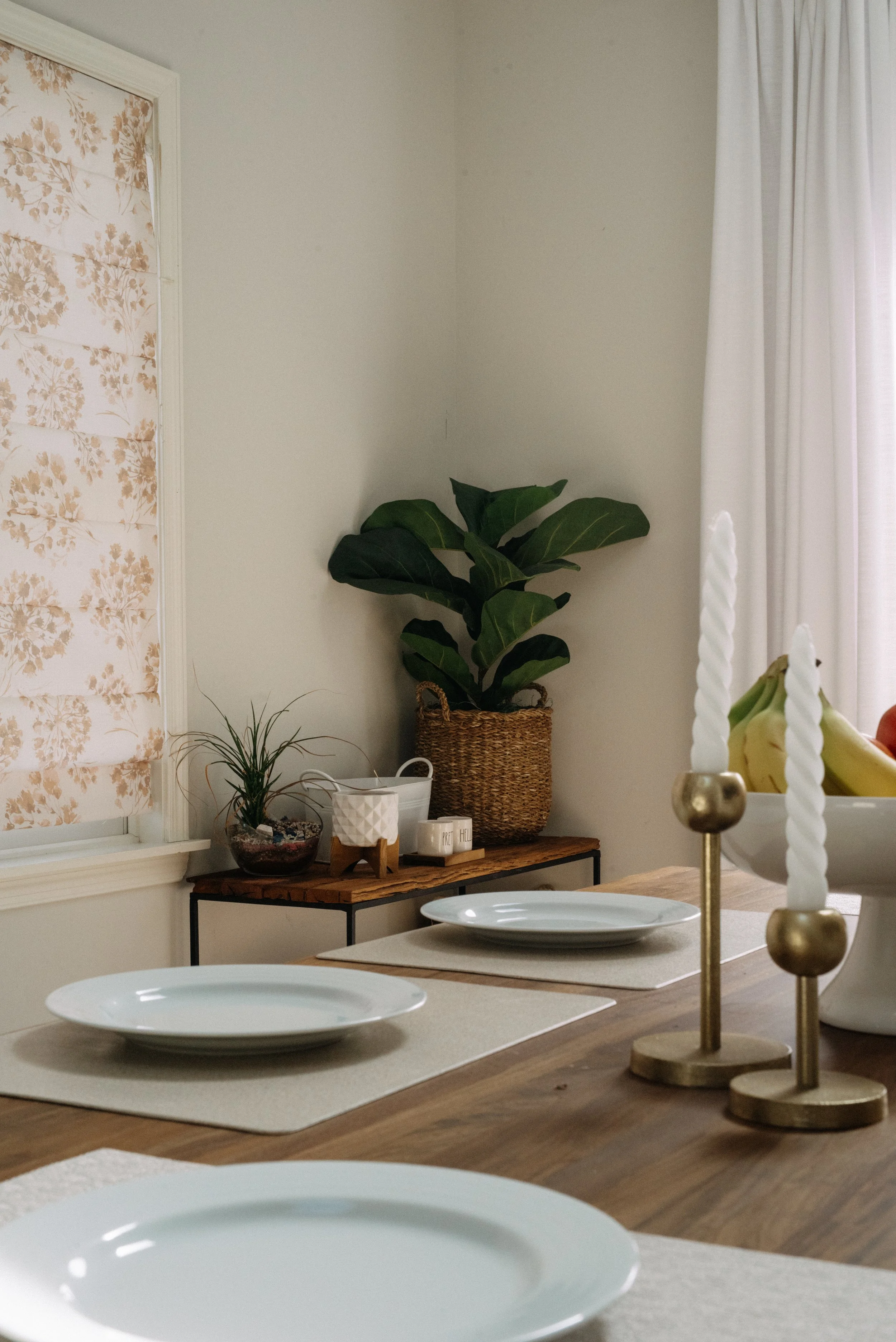 A dining table set with white plates, gold candleholders with white twisted candles, and a fruit bowl with bananas. A small black table against the wall holds a potted plant, a white container, a small white box, and a large potted plant in a woven b