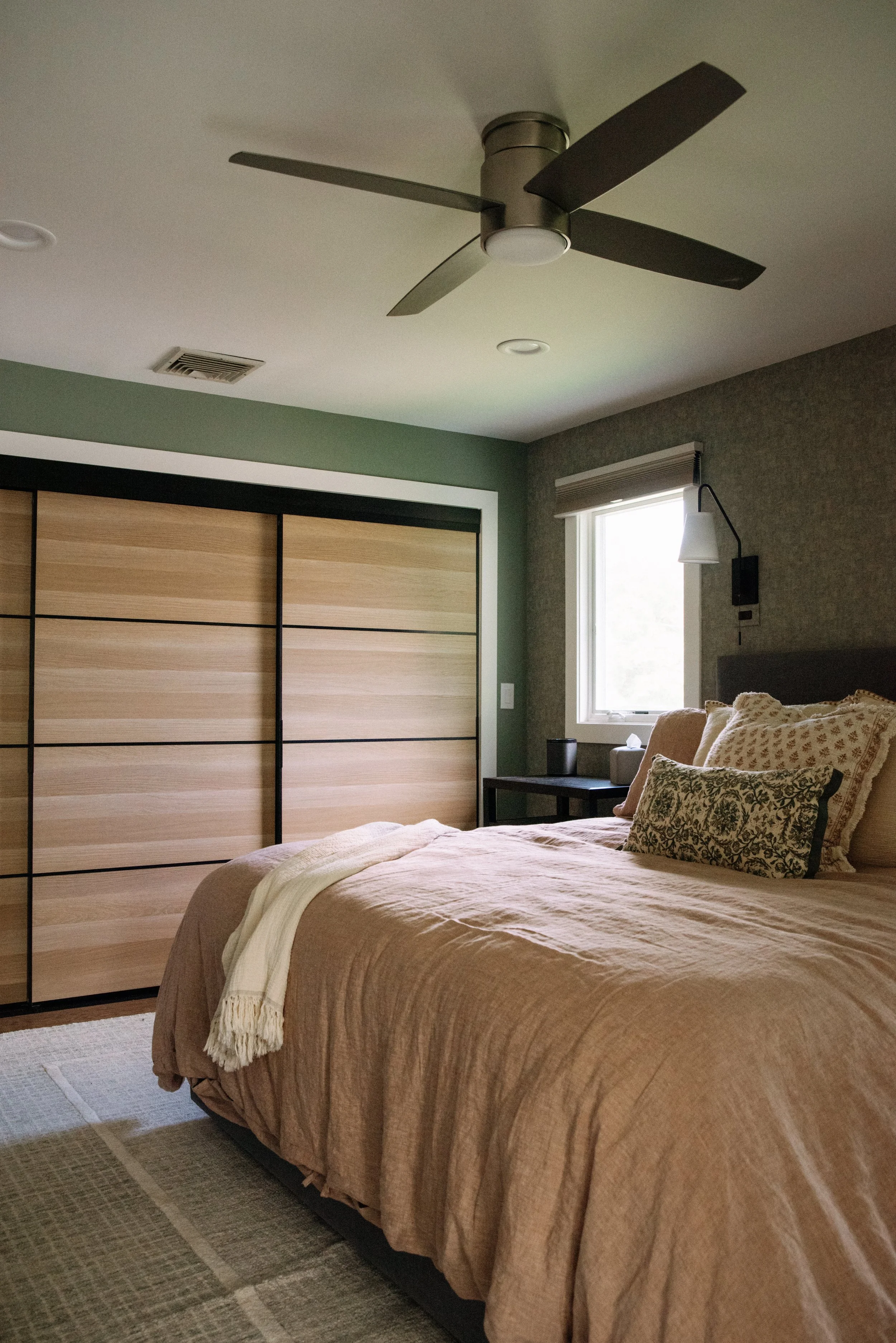 A bedroom with a bed, beige bedding, decorative pillows, a small window, a closet with sliding wood doors, and a ceiling fan.
