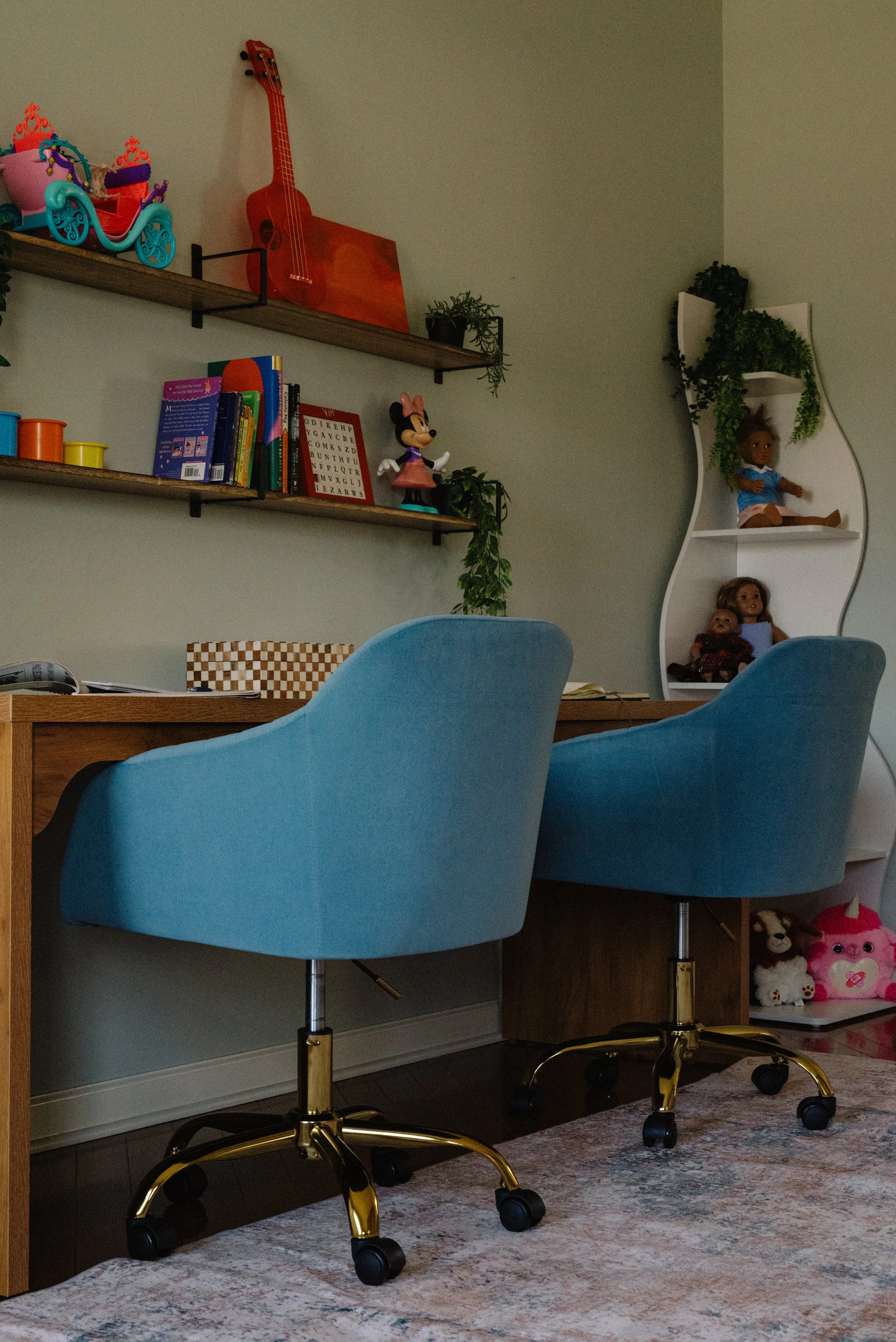 Home office or playroom with wooden desk, two blue chairs with gold bases, colorful bookshelf, toys, books, plants, and doll shelves.