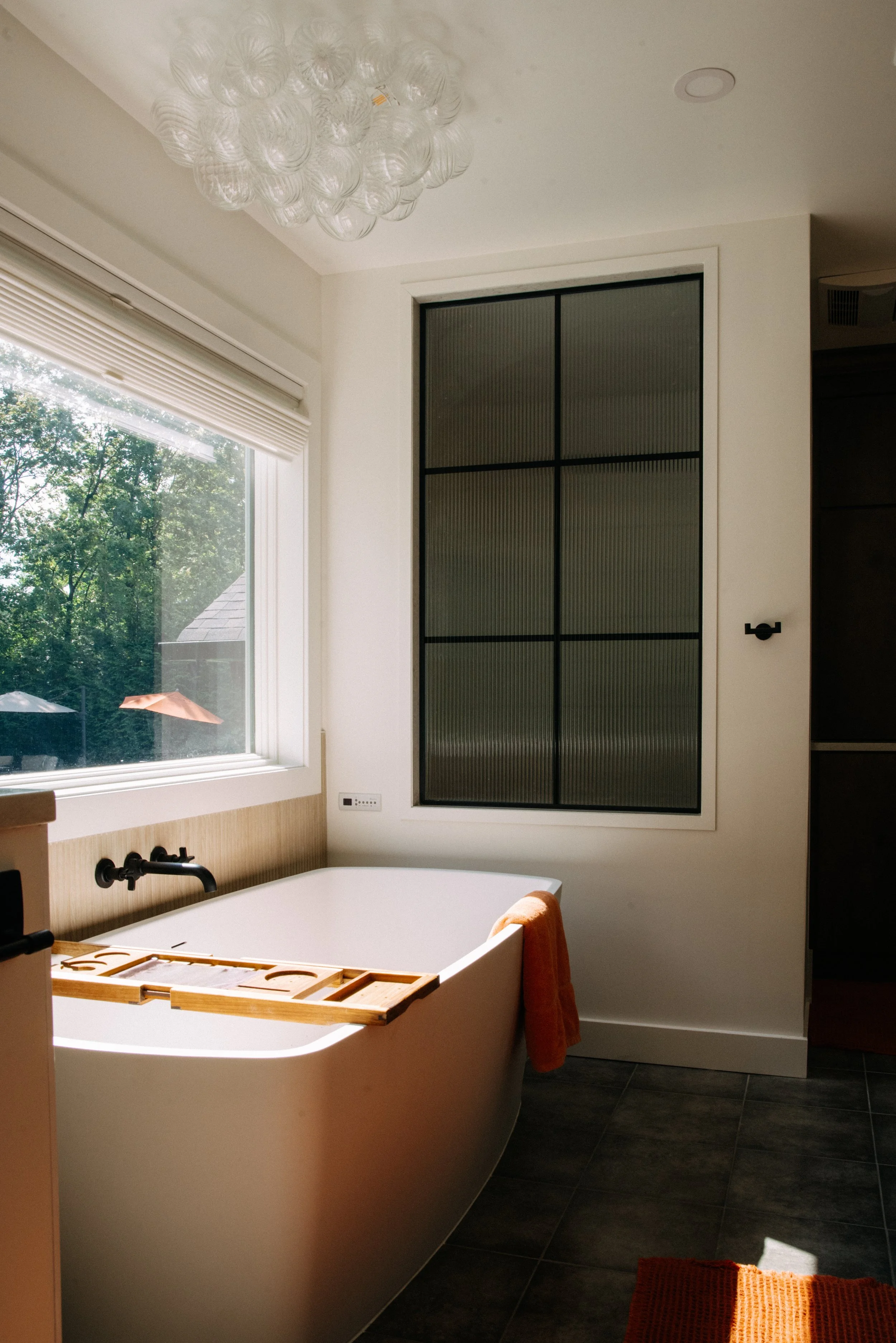Bathroom with a bathtub, orange towel hanging over edge, window with trees outside, and modern ceiling light fixture.