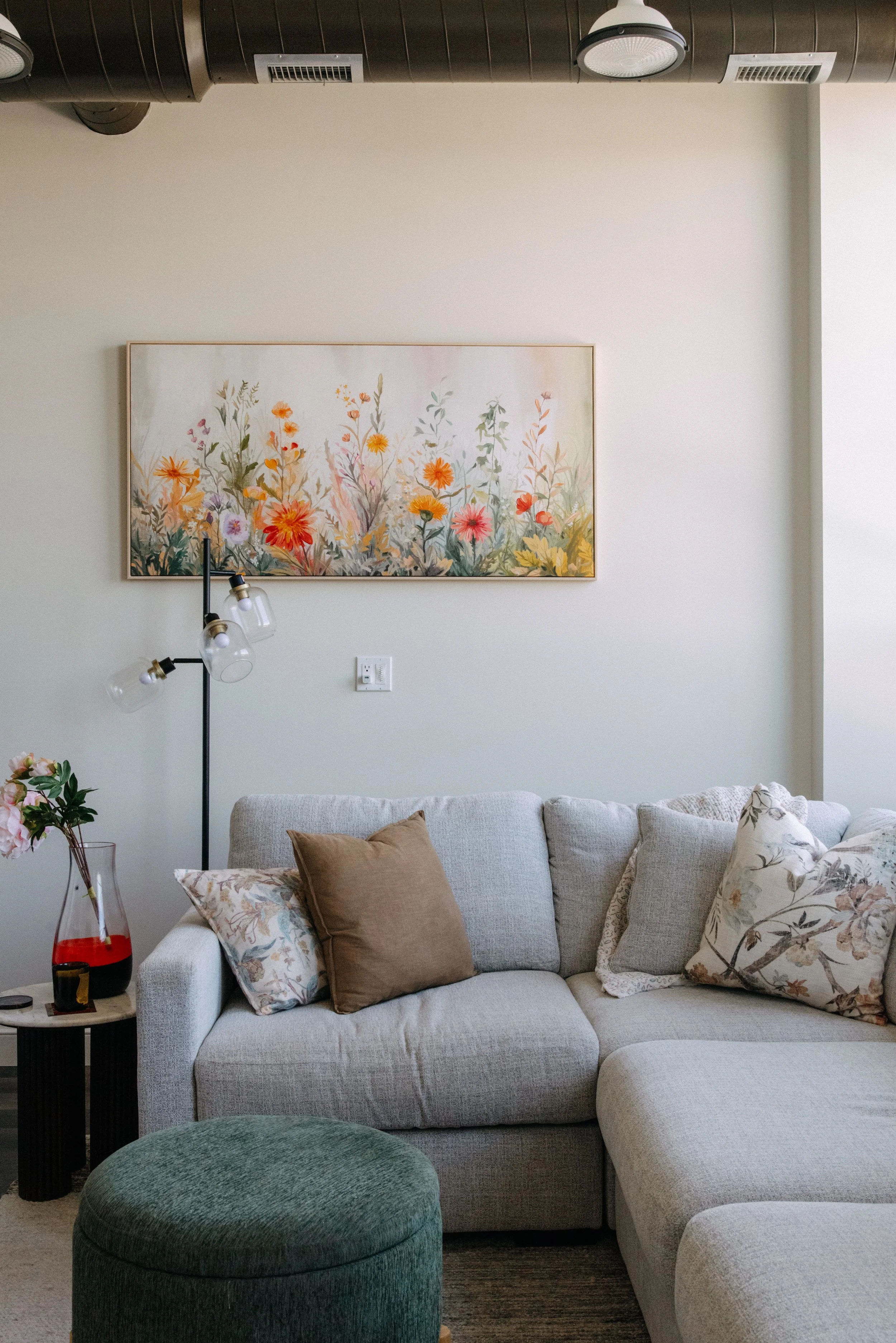 Living room corner with a light gray sectional sofa, decorative pillows, a small side table with a flower vase, a black floor lamp, a colorful floral painting on the wall, an overhead light, and visible air ducts.
