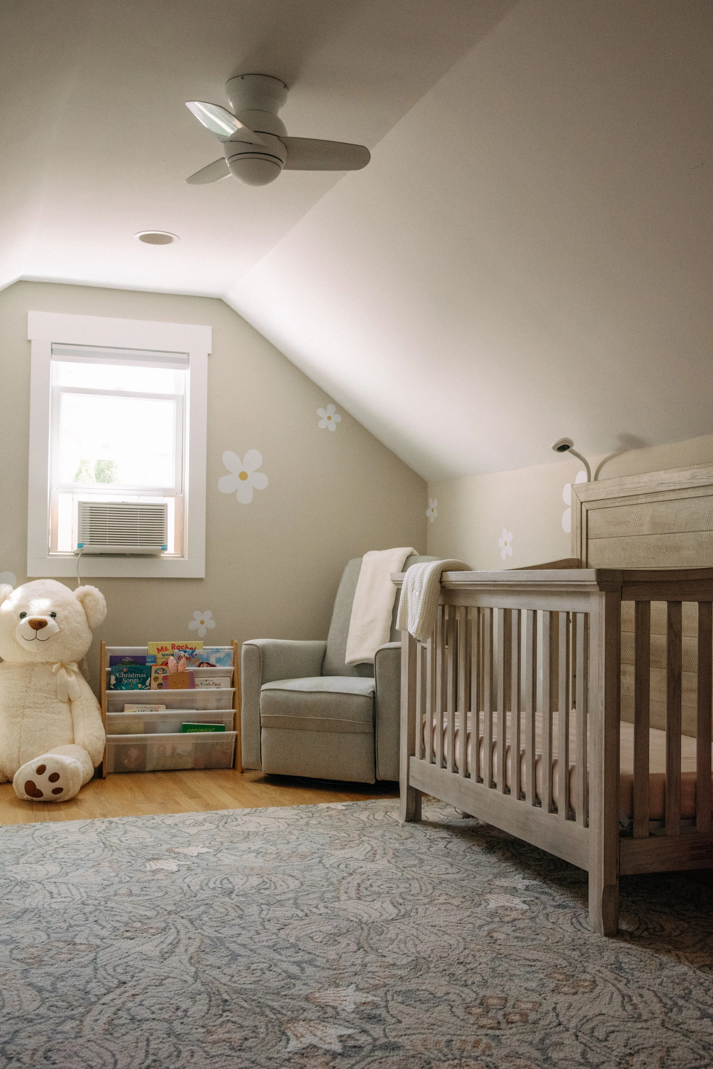 A cozy nursery with a crib, an armchair, a large teddy bear, a book cube, and wall decorations, featuring a slanted ceiling and a window with an air conditioning unit.