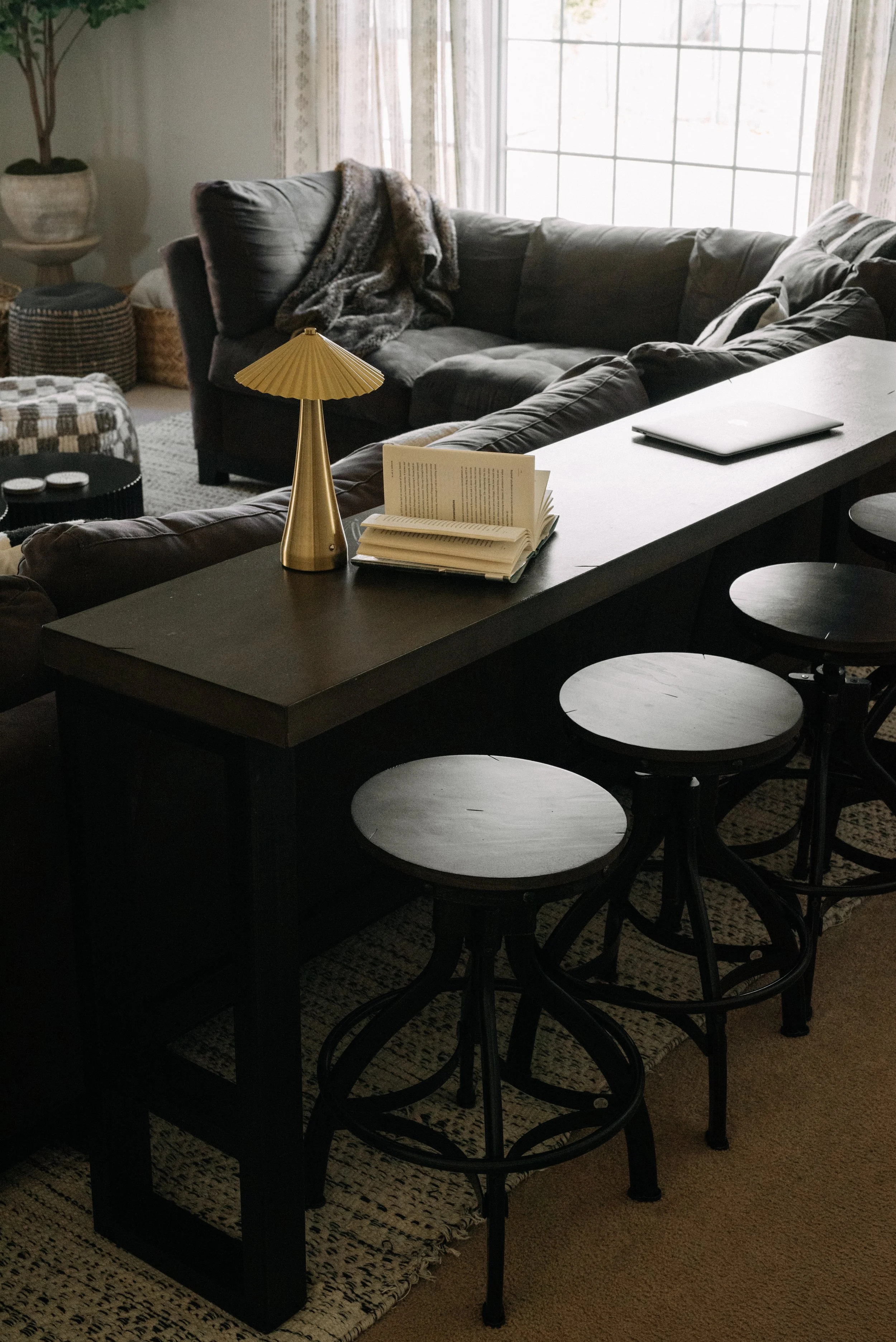 Living room with a black kitchen island, three black bar stools, a gray sofa with pillows, a blanket, a large window, a table lamp, an open book, and a laptop.
