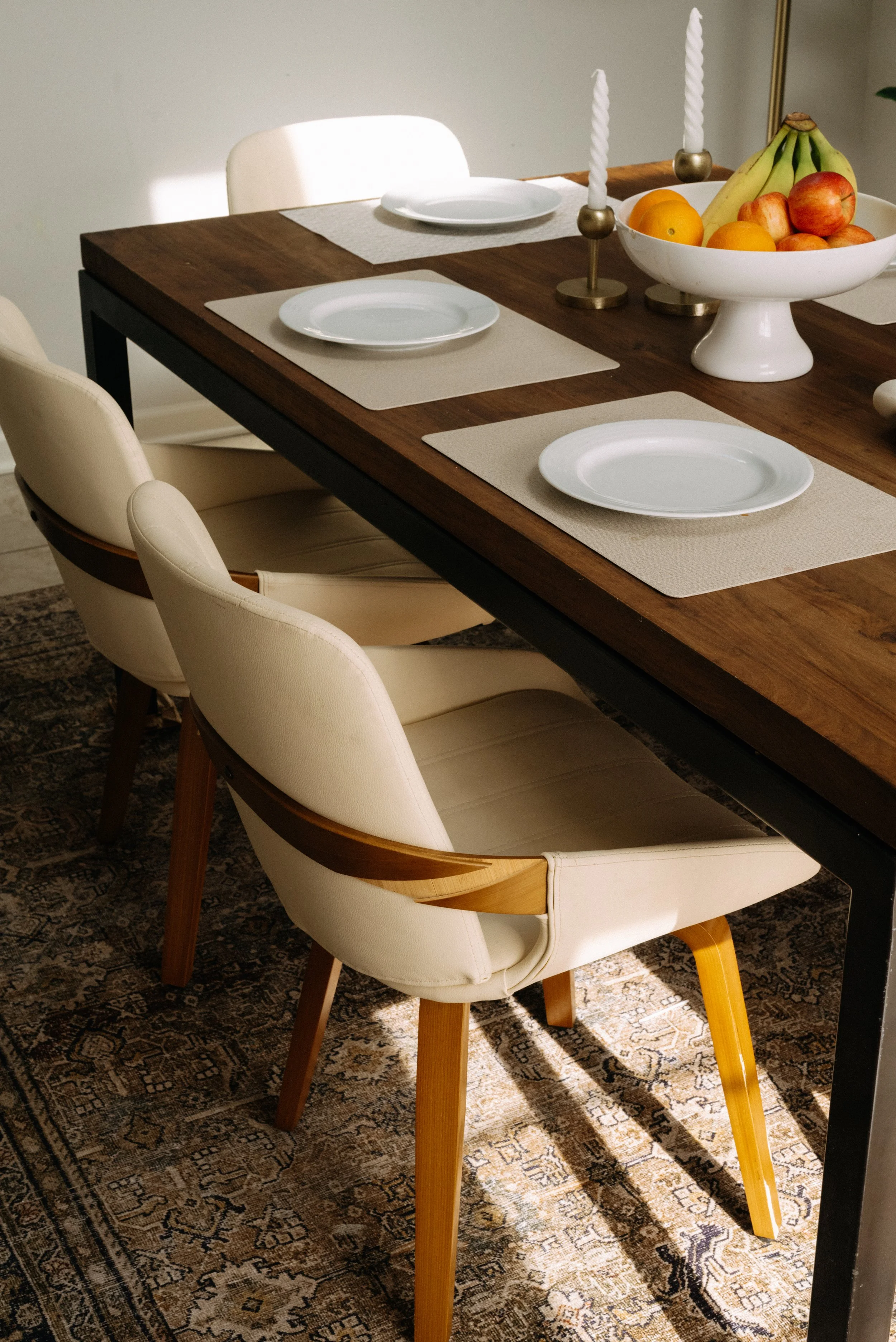 Dining table set with four cream-colored chairs, white plates, beige placemats, tall white candles in holders, and a bowl of assorted fruits including bananas, oranges, and apples. The table has a wooden surface and the setting is on a patterned rug.