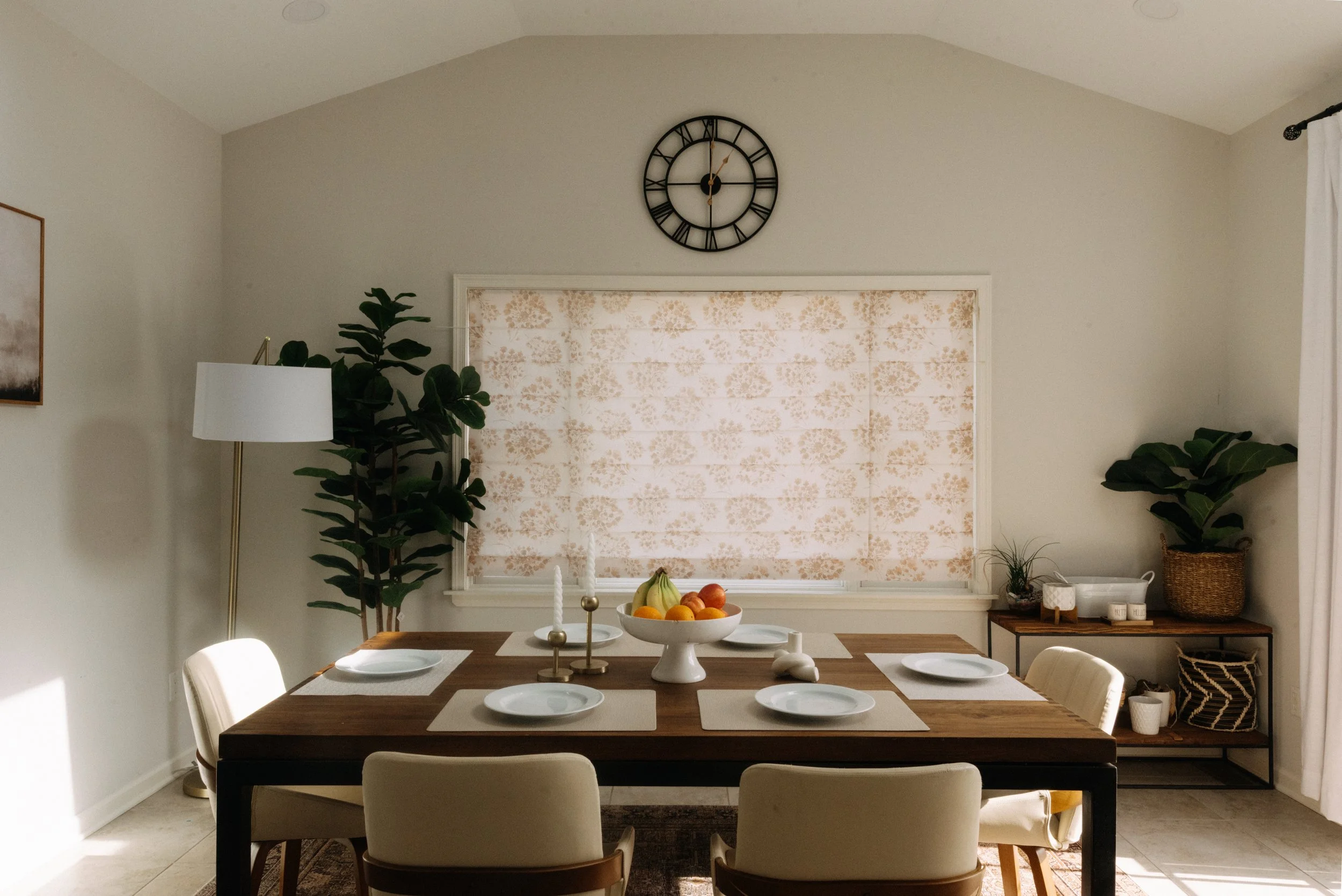 A dining room with a wooden table set for five, featuring white plates, a bowl of fruit, white candles, and surrounded by beige chairs. A large clock is on the wall above a white window with patterned curtains. There are two large potted plants and a