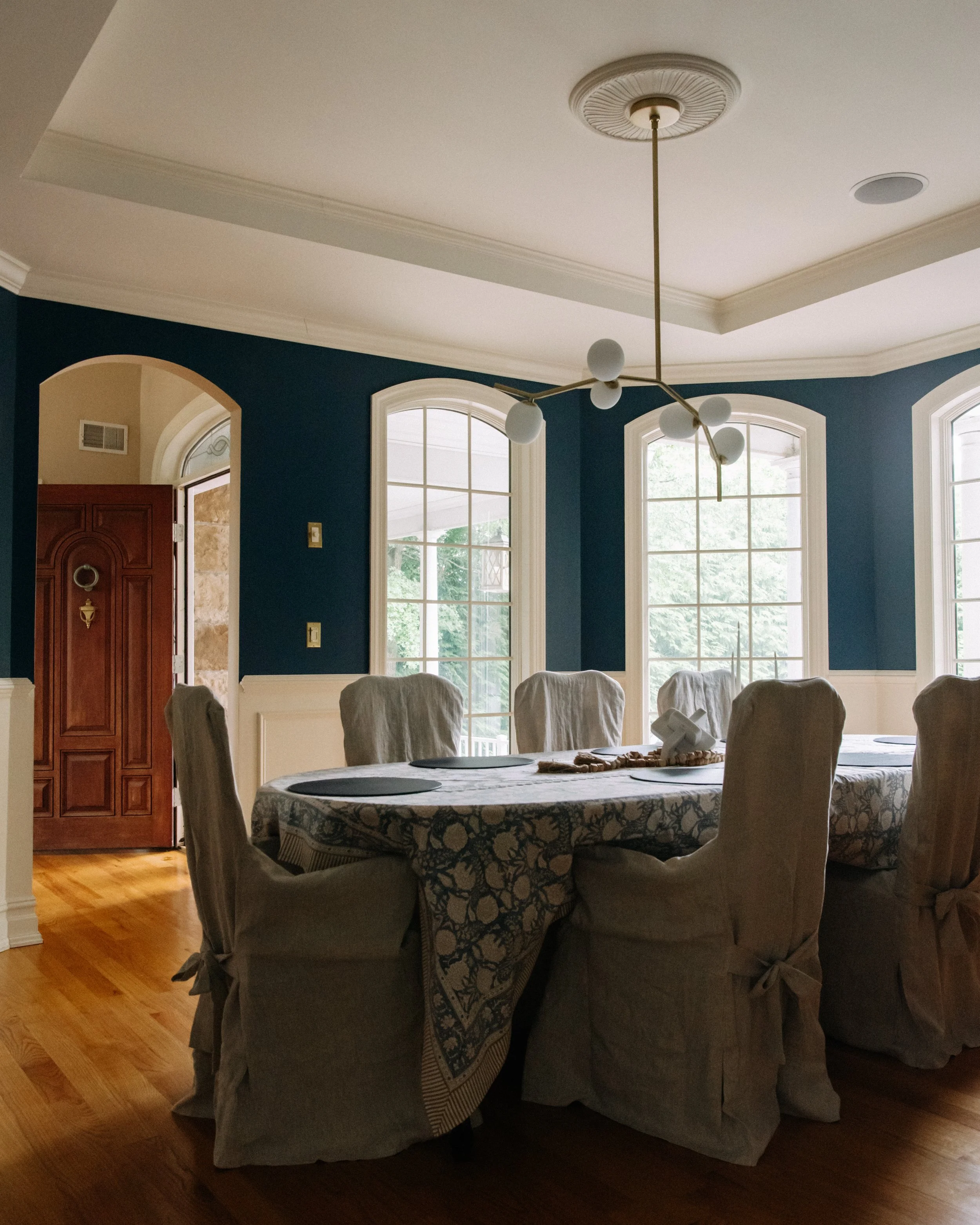 A dining room with a round table covered with a tablecloth, surrounded by eight chairs with slipcovers tied at the back. The room has dark blue walls, large windows allowing natural light, and a modern chandelier hanging from the ceiling. There is a