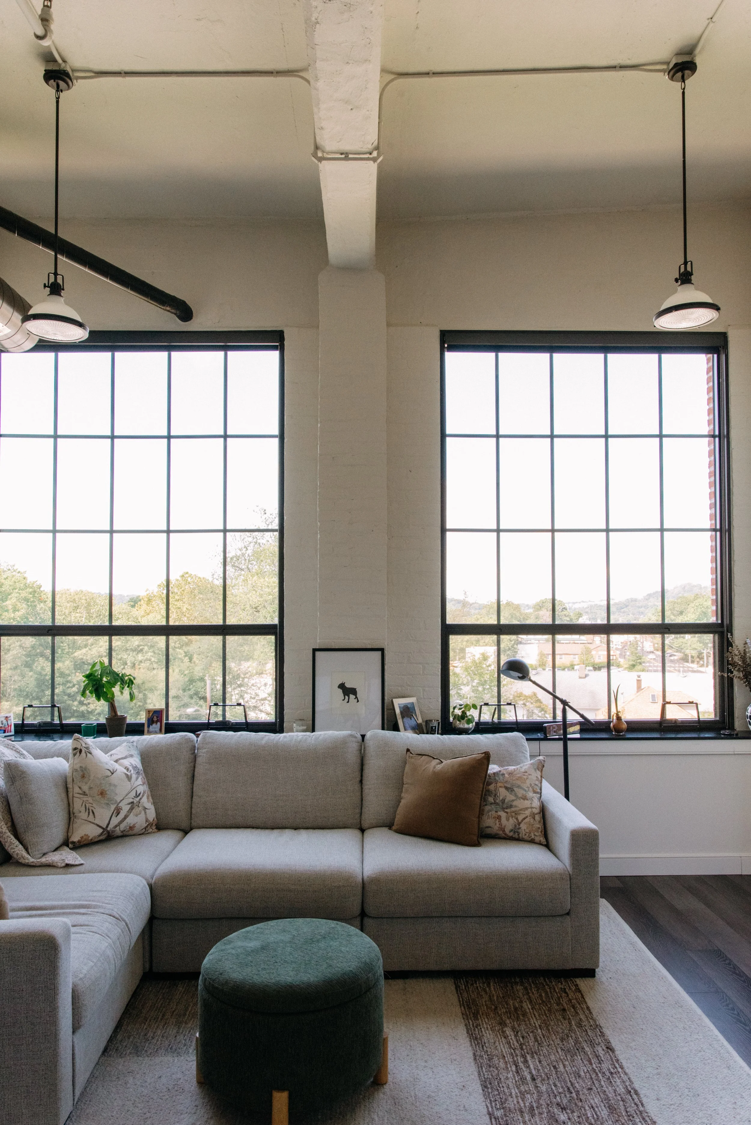Living room with large windows, a beige sectional sofa with patterned and solid pillows, a green ottoman, and decorated ledges beneath the windows.
