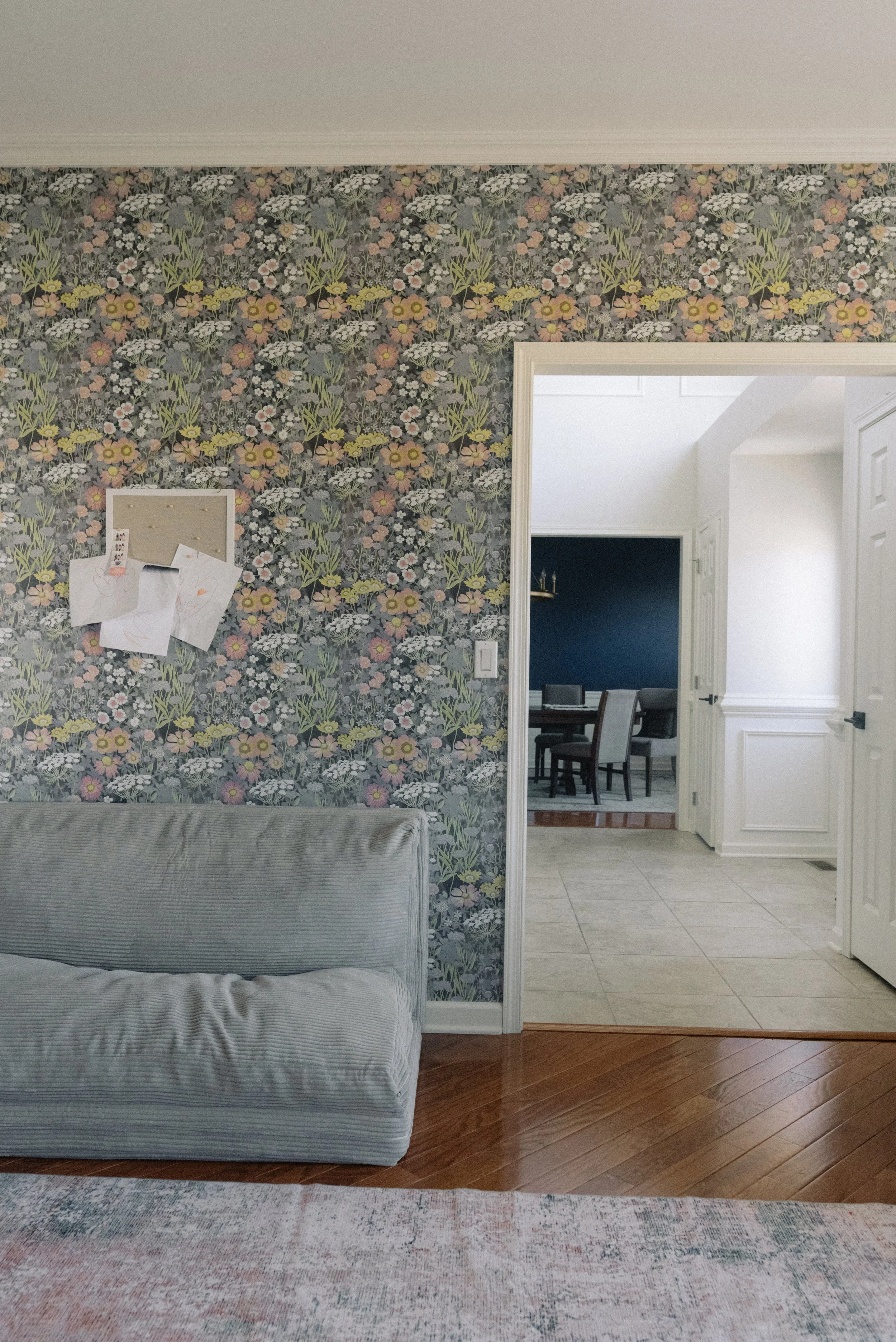A living room with a floral-patterned wallpaper, a gray armchair, a corkboard with papers, and a doorway leading to a dining room with a dark blue wall and a dining table with chairs.