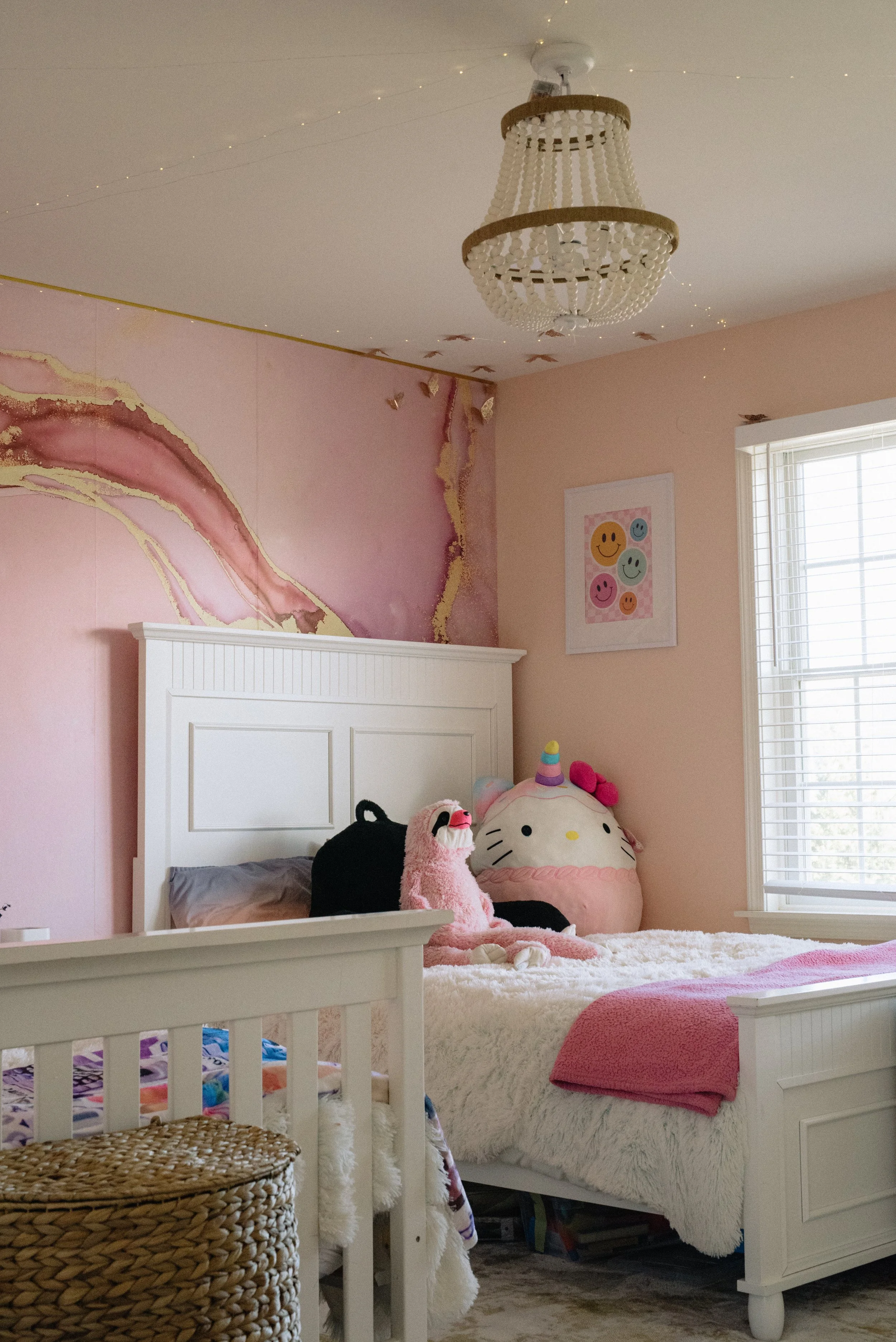 A cozy children's bedroom decorated with pink and white furniture, plush toys including a Hello Kitty pillow, a stuffed sloth, and a unicorn plush, soft blankets, and wall art with smiley faces. The room features a window with blinds, a chandelier, a