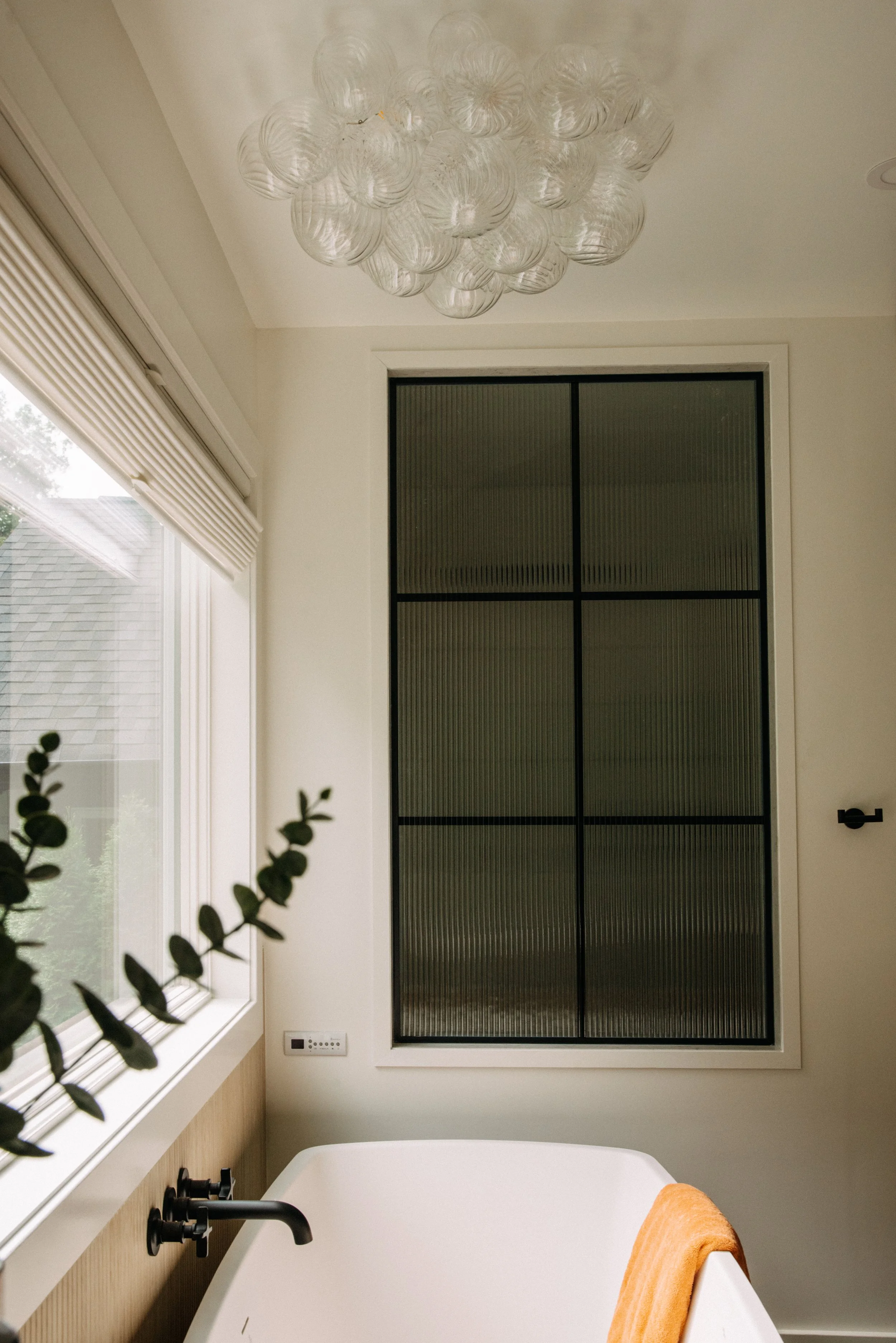 A modern bathroom featuring a white bathtub with a yellow towel draped over its edge, a large window with blinds, a black-framed textured glass window, a contemporary black faucet, a wooden panel behind the tub, a small control panel, and a decorativ