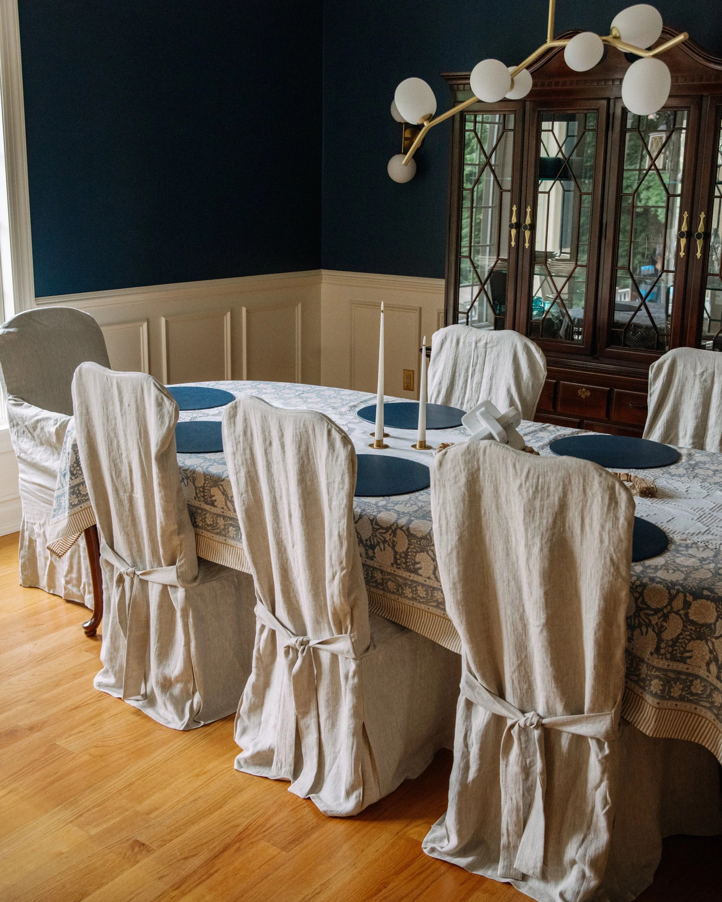 A dining room with a wooden table covered in a patterned tablecloth, six dining chairs with white slipcovers tied at the back, a dark blue wall, a wooden china cabinet with glass doors, and a modern chandelier with globe lights.