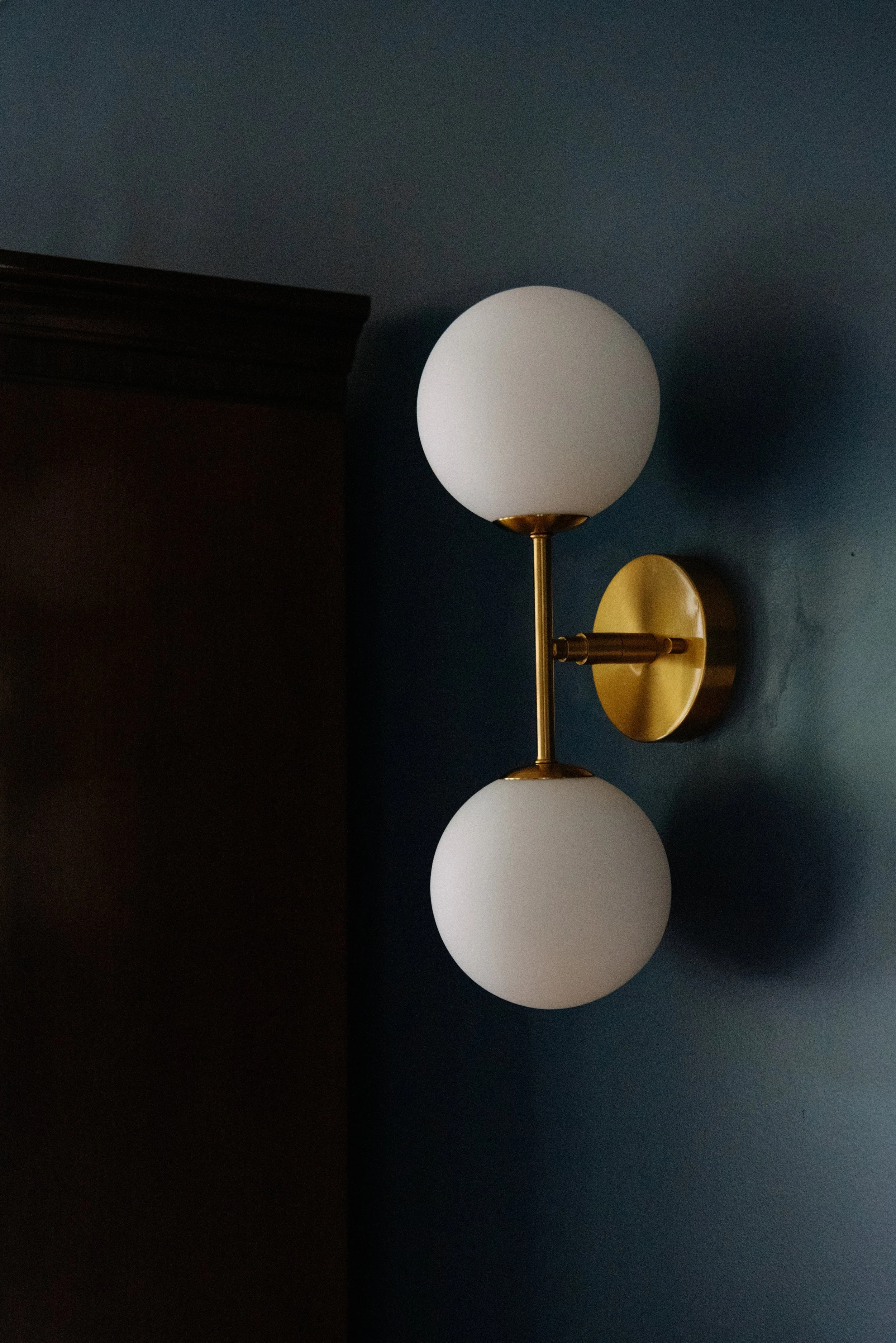 A modern wall sconce with two white globe lights mounted on a gold-colored fixture attached to a dark blue wall.