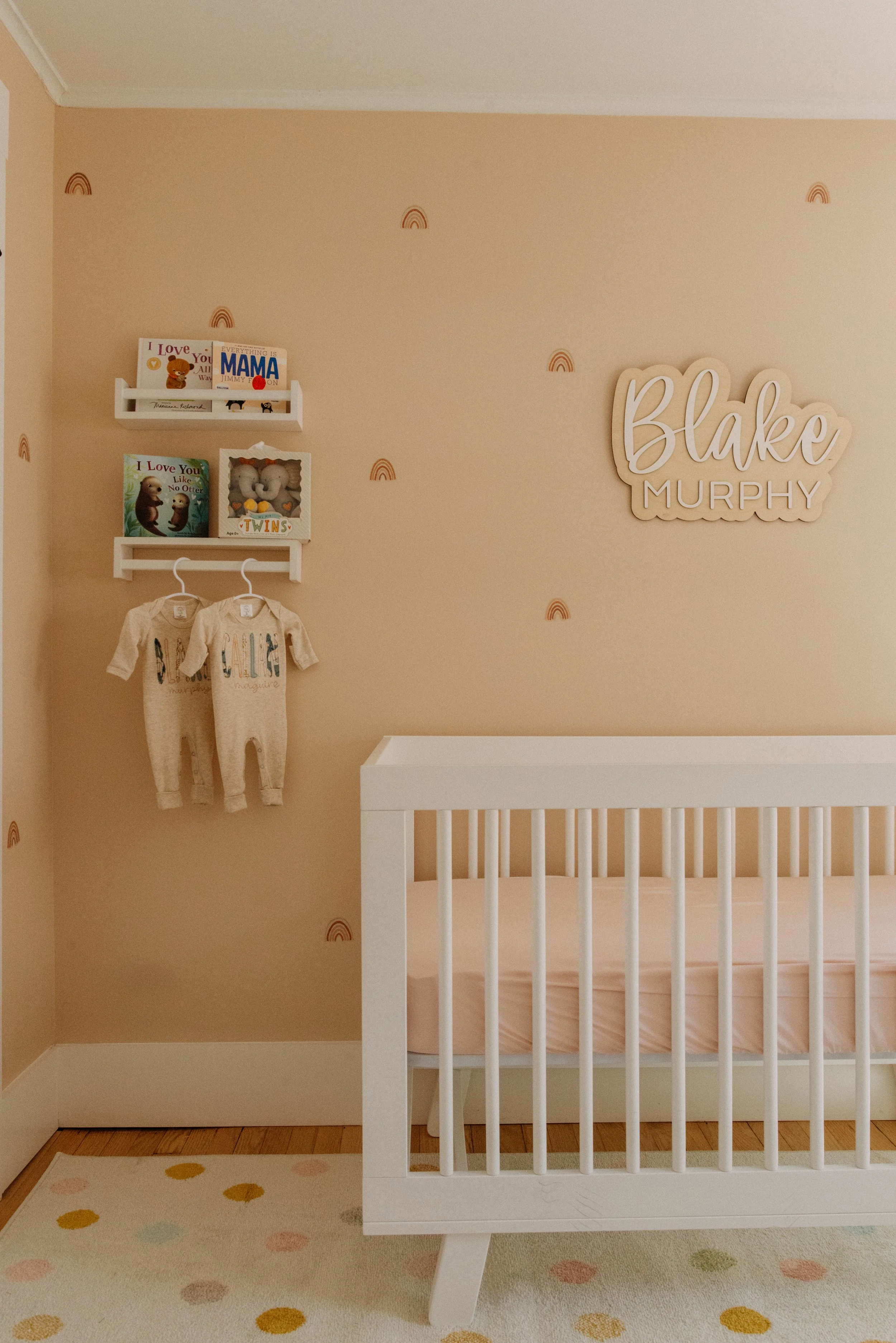 Nursery room with beige walls decorated with small rainbow decals. There is a white crib and two beige baby onesies hanging on hooks. Two shelves hold children's books and stuffed animals. A wooden name sign reads 'Blake Murphy.' The floor has a polk