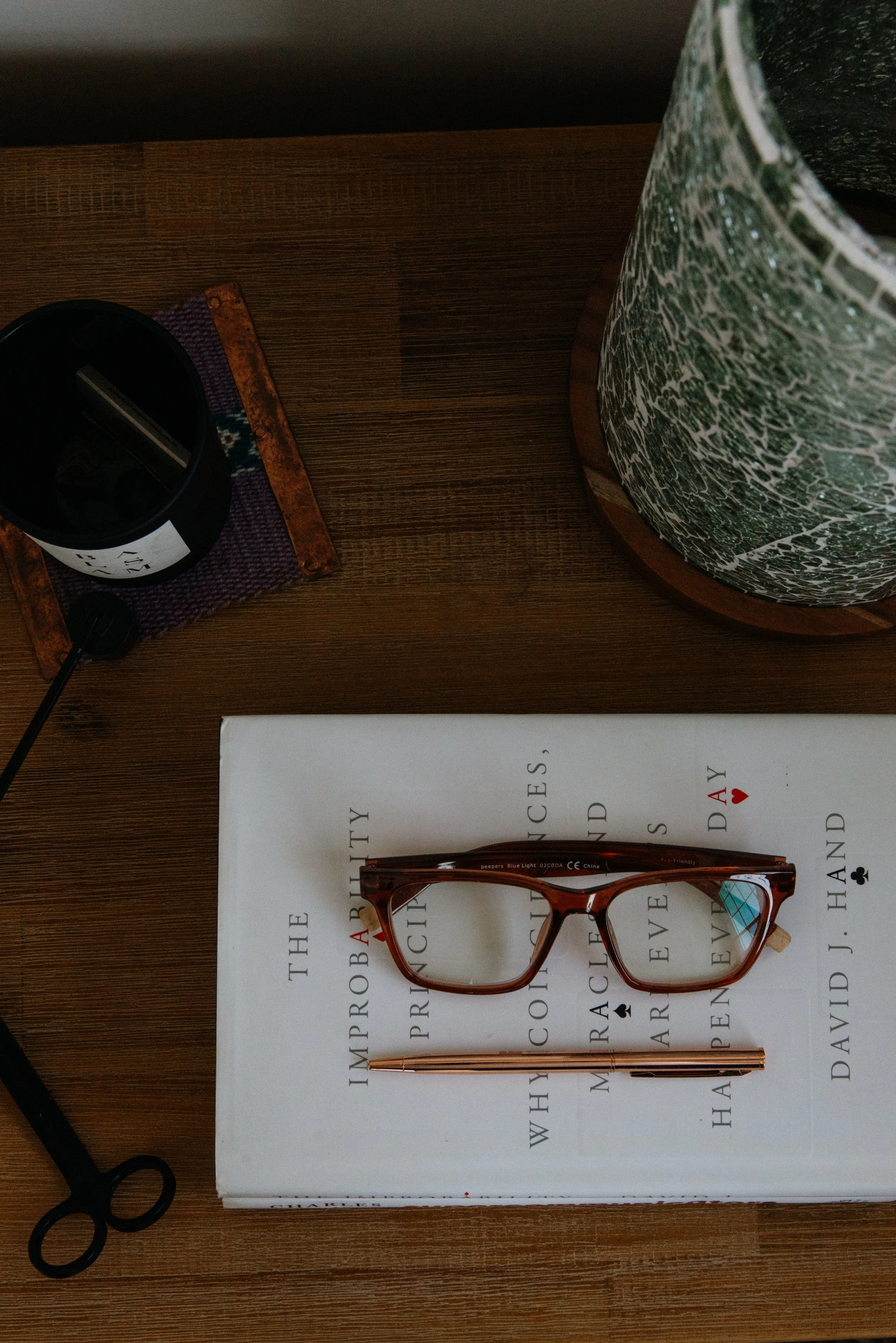 A pair of brown glasses and a gold pen are placed on top of a book with the author's name, David J. Hanny, on a wooden desk. To the right, there is a large green and white patterned ceramic vase, and to the left, a black candle holder with a lit cand