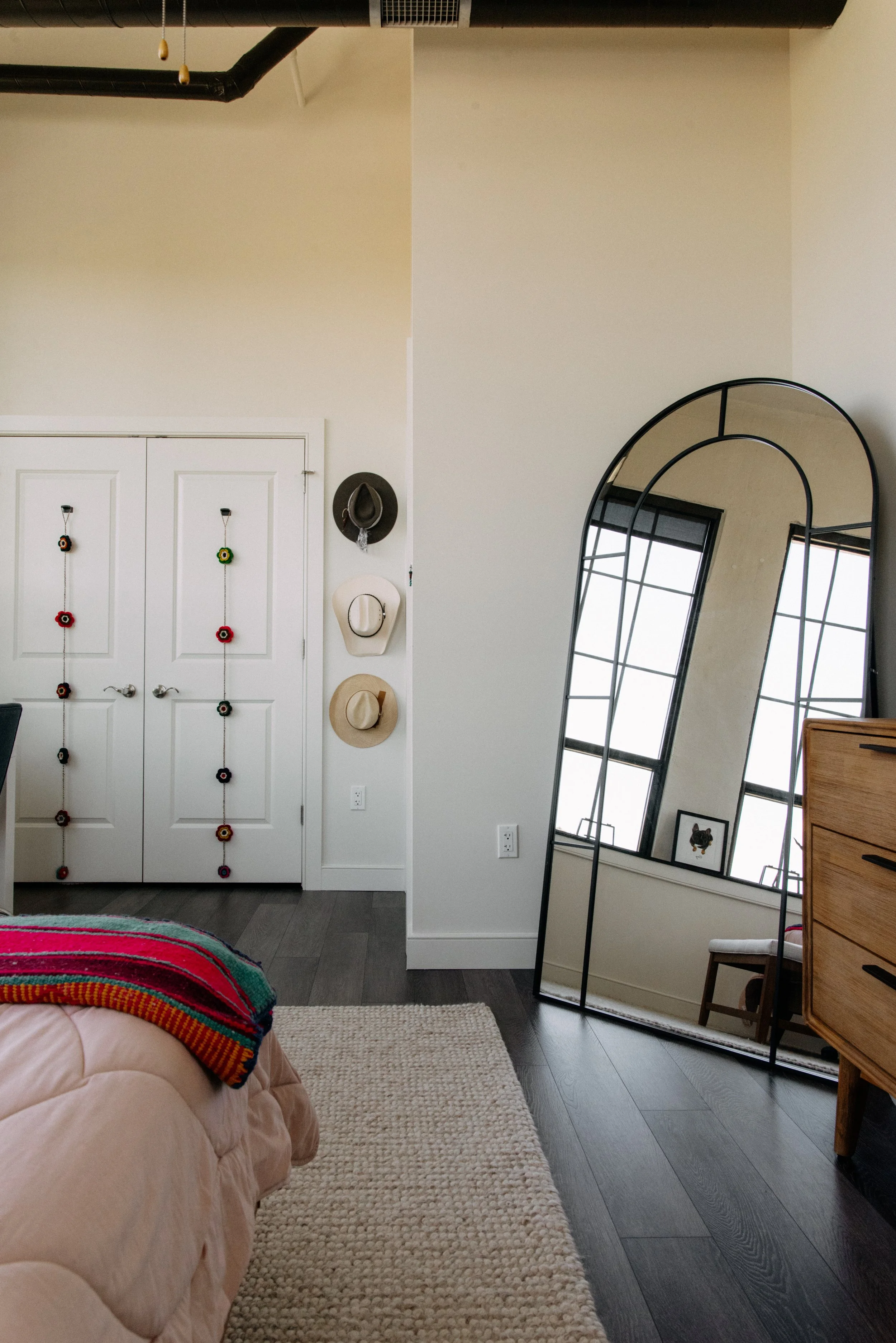 A bedroom corner with a large, arched, black-framed mirror leaning against the wall near a window. There is a dresser with wooden drawers to the right of the mirror, and a small framed picture of a dog on the windowsill. On the left side, there is a
