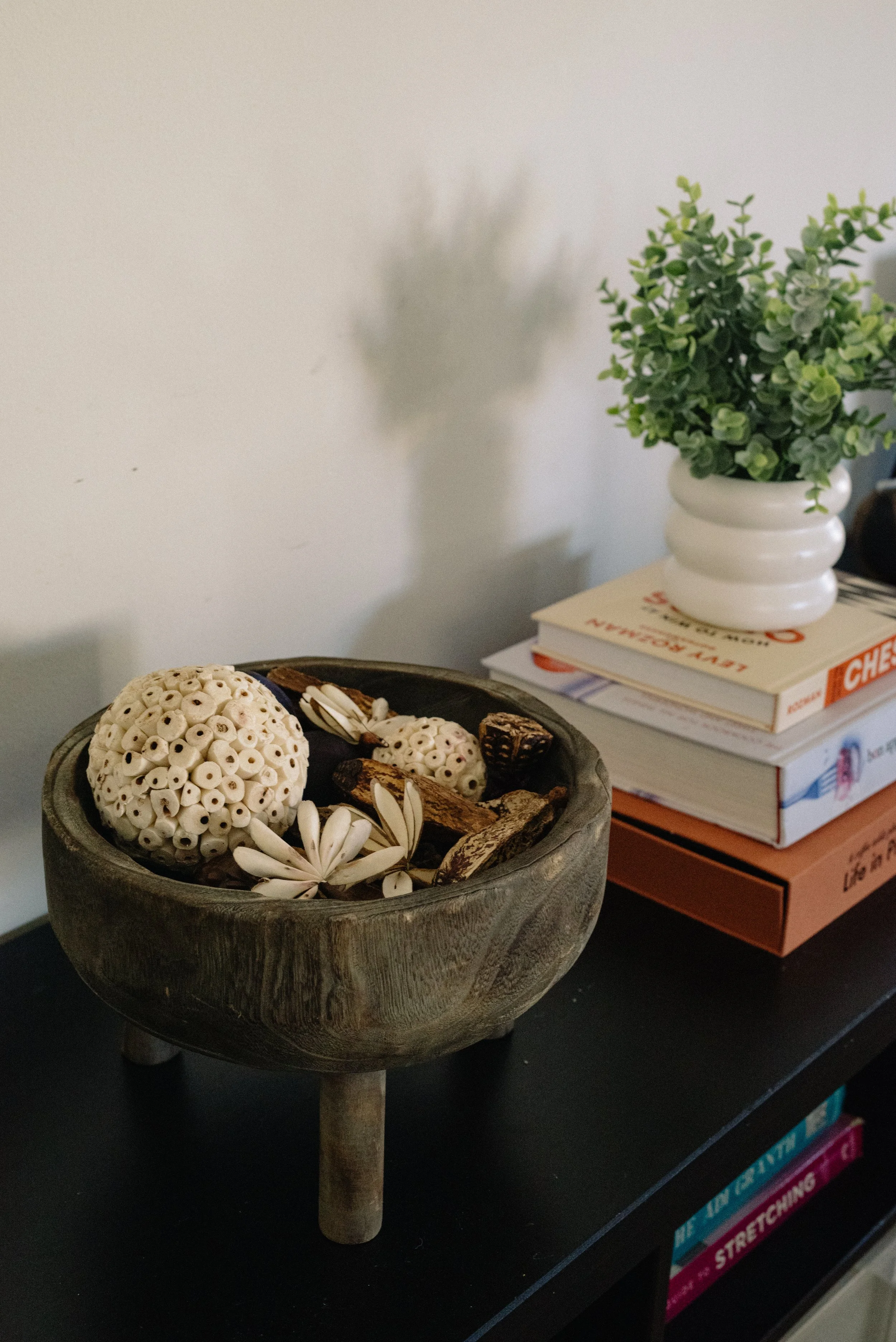 Decorative bowl with dried tan and white floral and seed pods on a black table next to a stack of books with a potted green plant.