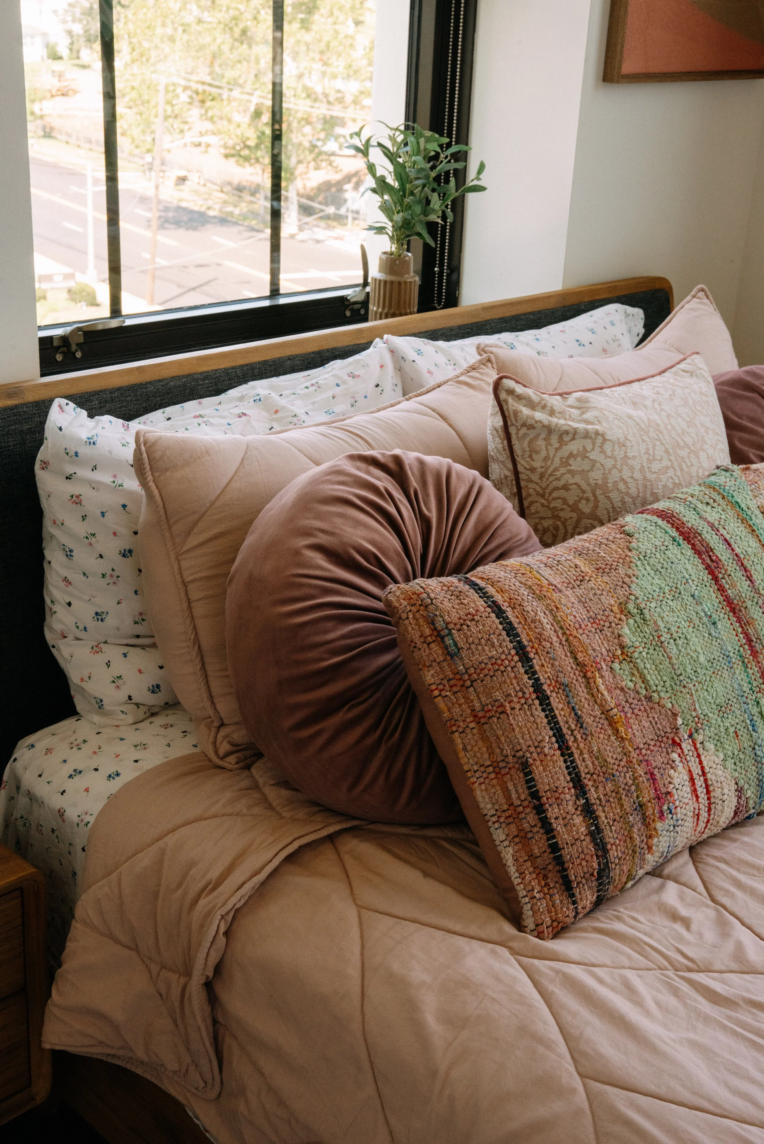A cozy bedroom bed with multiple decorative pillows, blankets, and a window with a small plant on the windowsill.