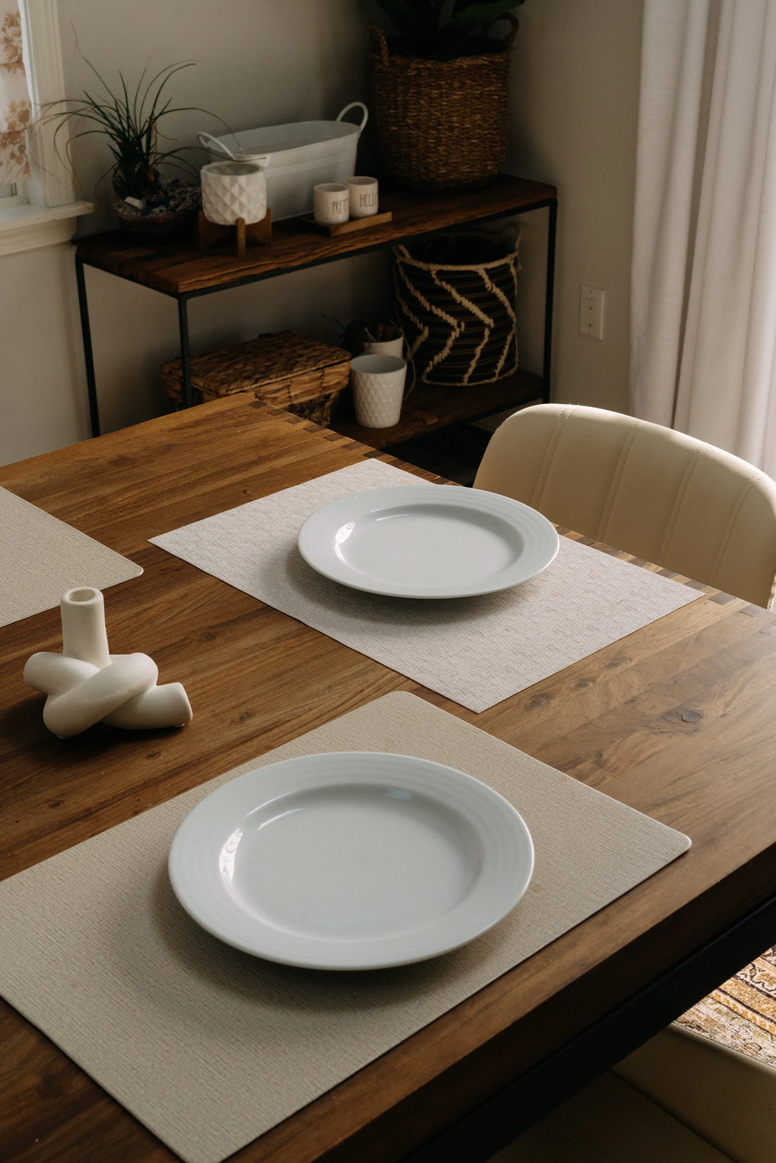 Dining table set with two white plates on beige placemats, a white decorative vase, and a wooden table with a natural finish. In the background, a wooden sideboard with baskets, potted plants, and mugs, with a window and curtains to the right.