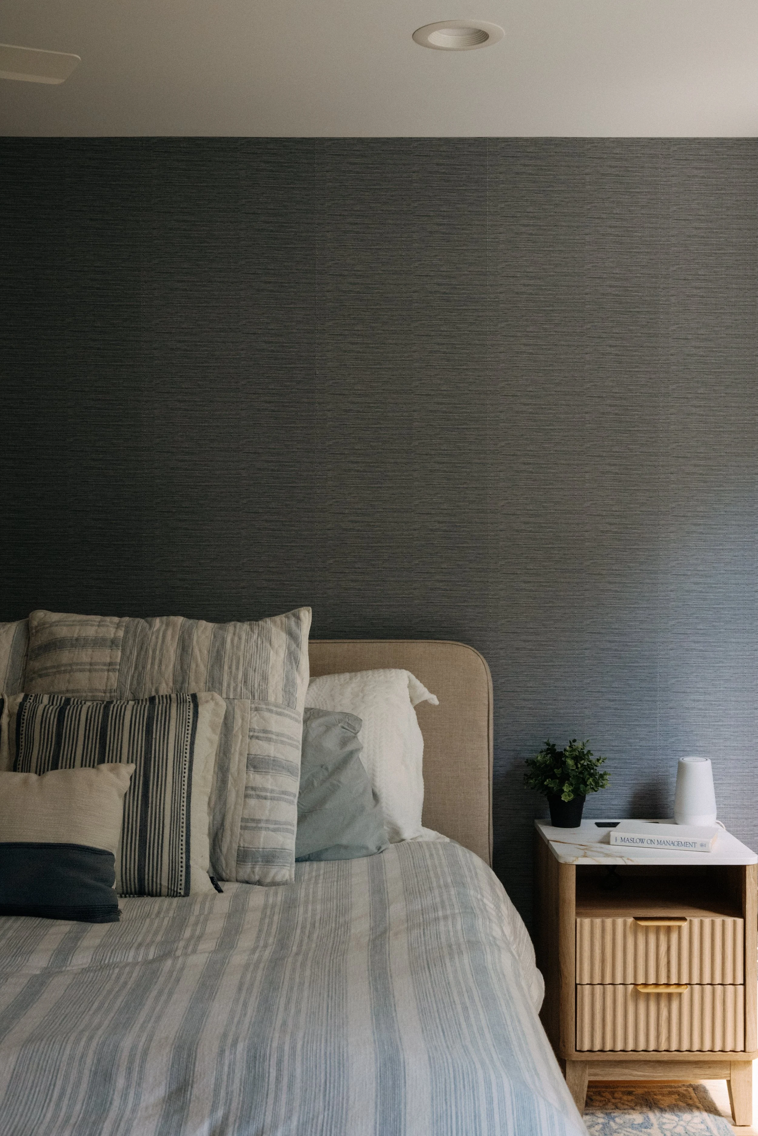 A neatly made bed with multiple pillows and striped bedding, beside a wooden nightstand with a lamp, a plant, and a book, against a textured dark gray wall.