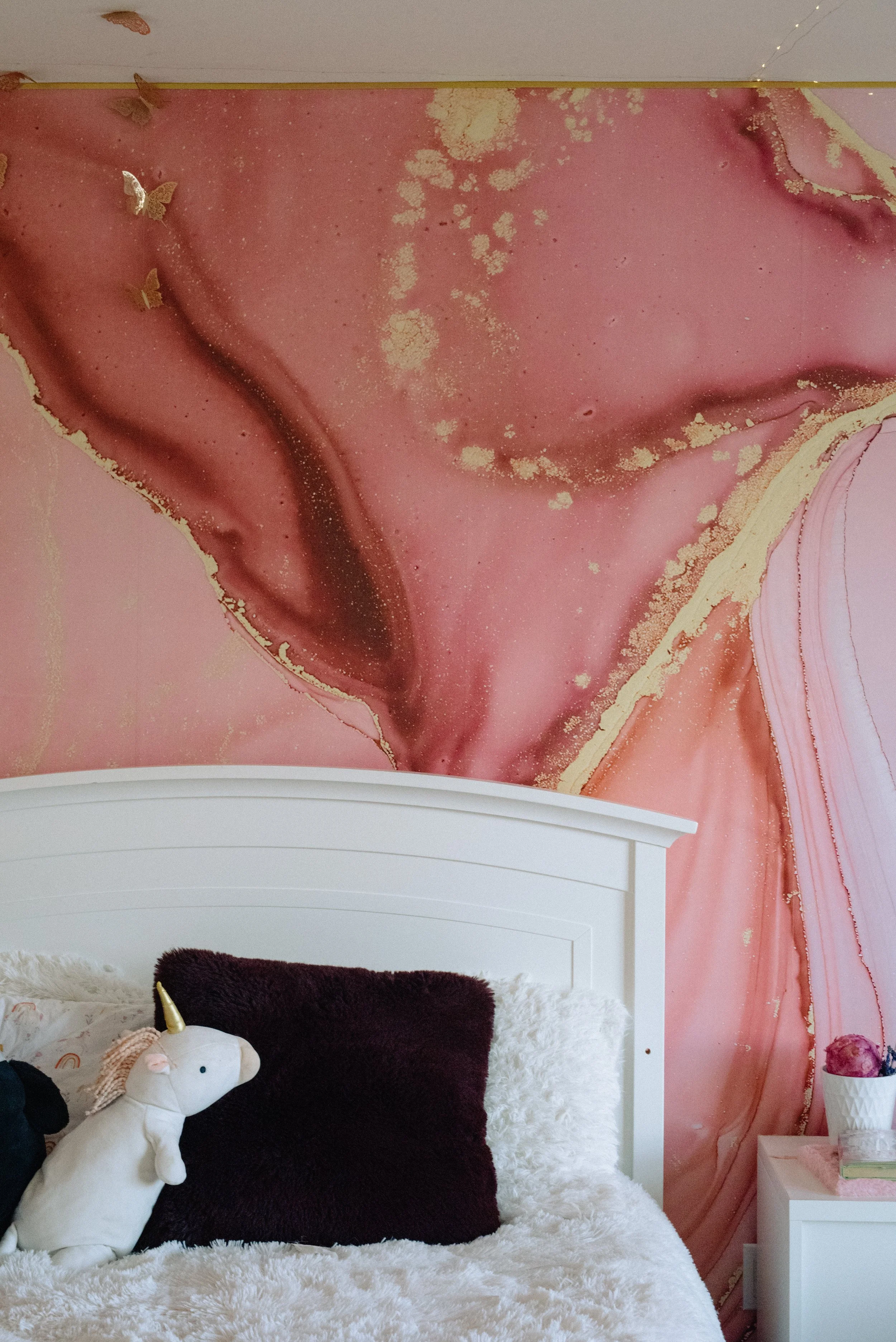 A bed with a white headboard, fluffy white and dark purple pillows, a plush unicorn toy, and a pink bedside table with flowers, against a pink and gold abstract mural wall.