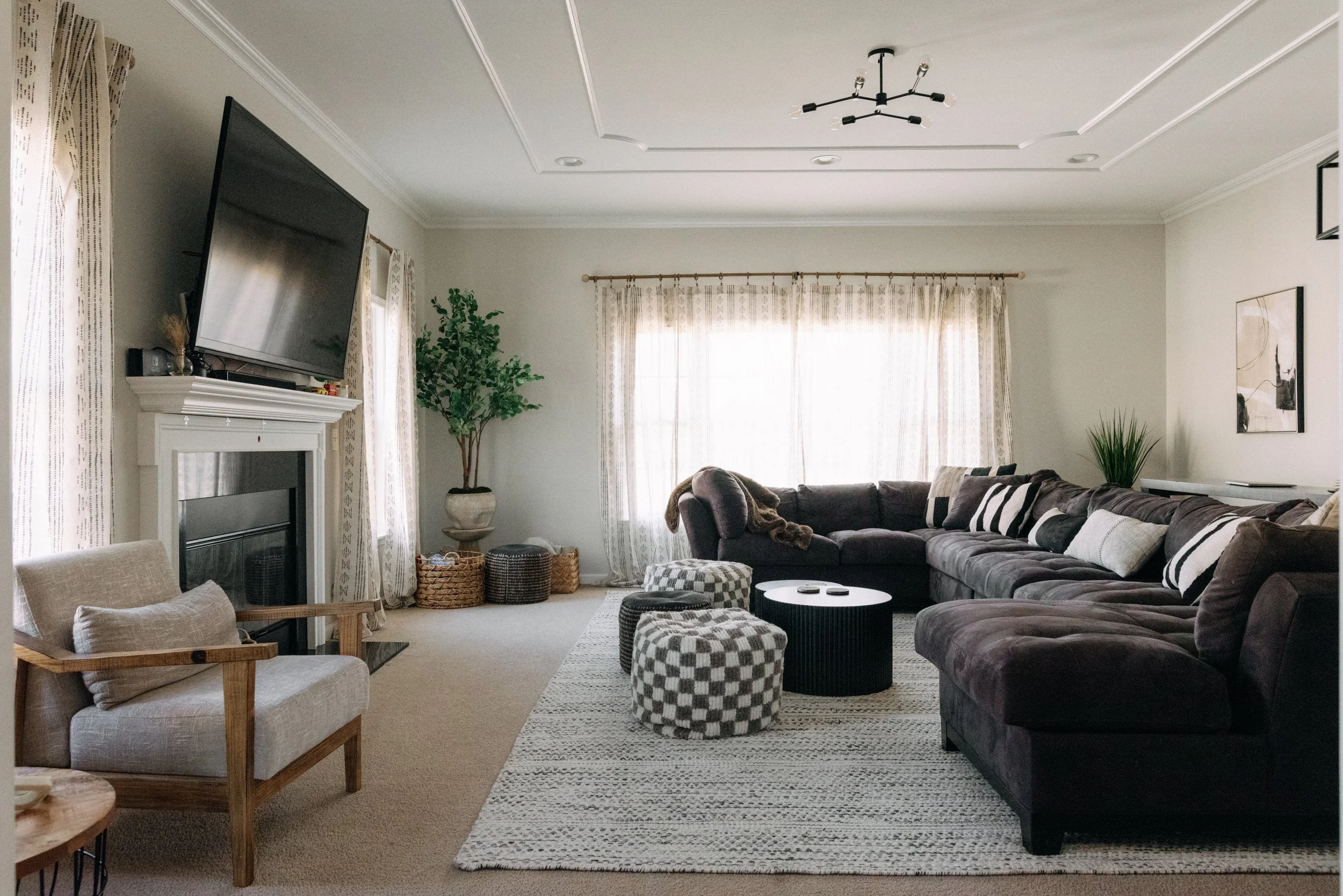 Living room with a large L-shaped dark sofa, patterned pillows, a coffee table, armchair, potted plants, curtains, and a mounted flat-screen TV above a fireplace.