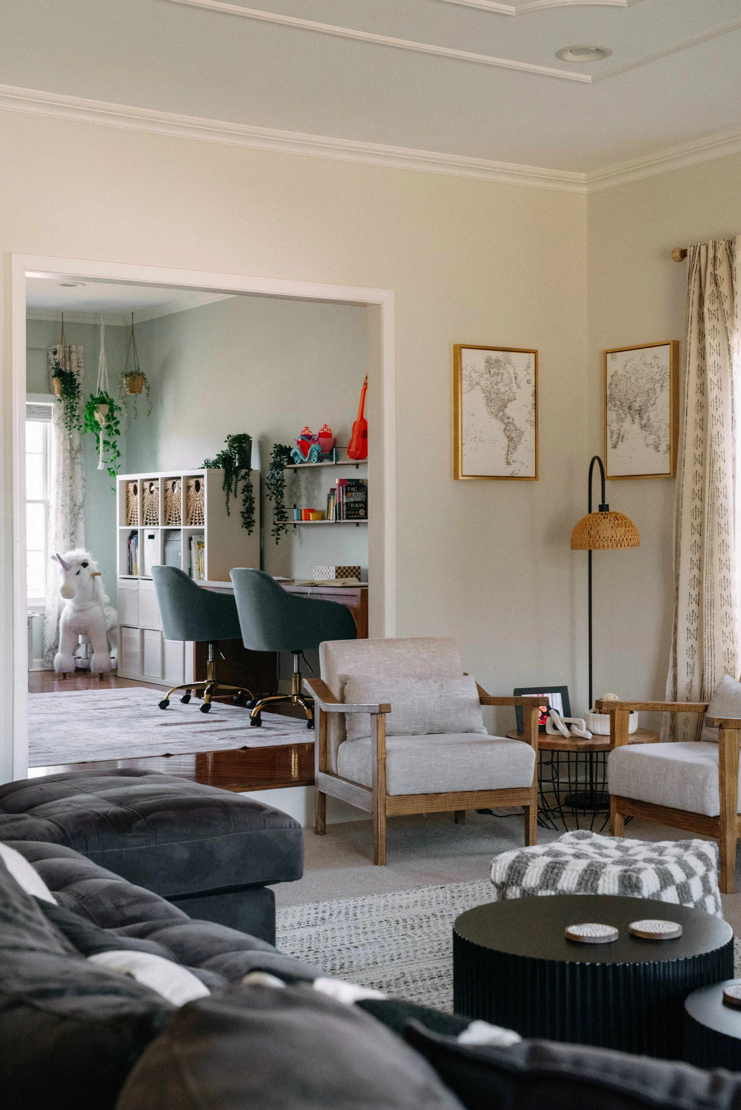 Living room with beige armchairs, a black coffee table, and wallpapered curtains. Through an open doorway, a room with a white unicorn statue and shelves with books and toys can be seen.