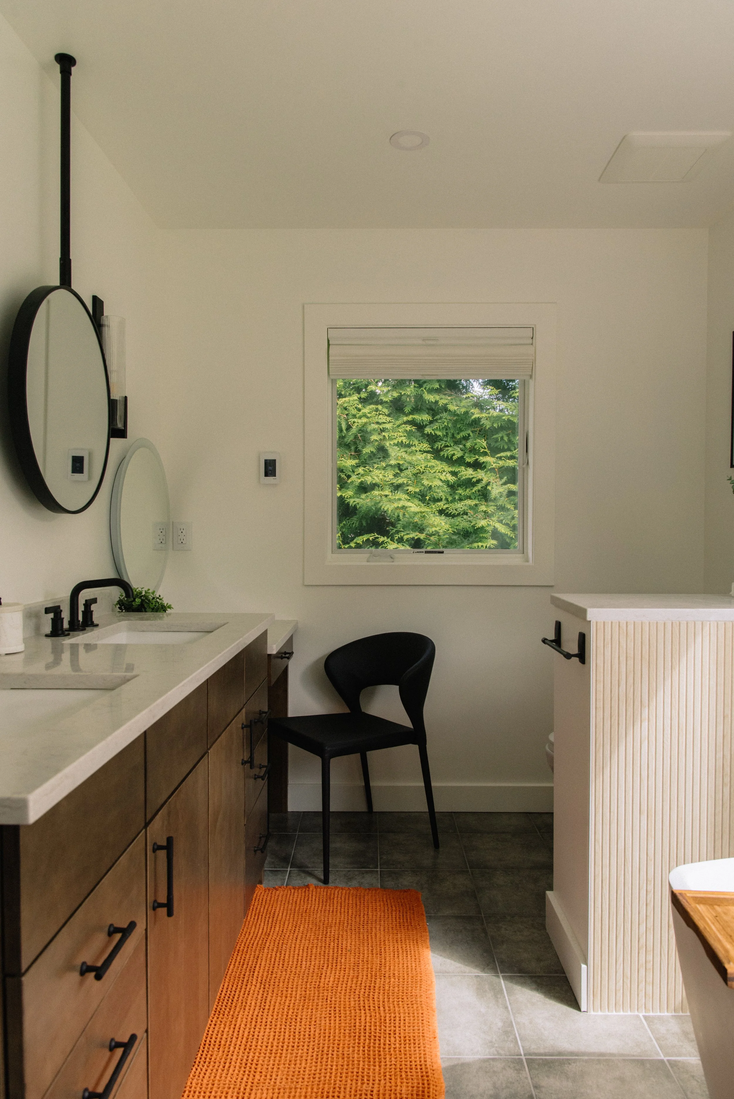 Modern bathroom with double sinks, black fixtures, wooden cabinets, a window with greenery outside, a black chair, an orange rug, and a partial view of a toilet.