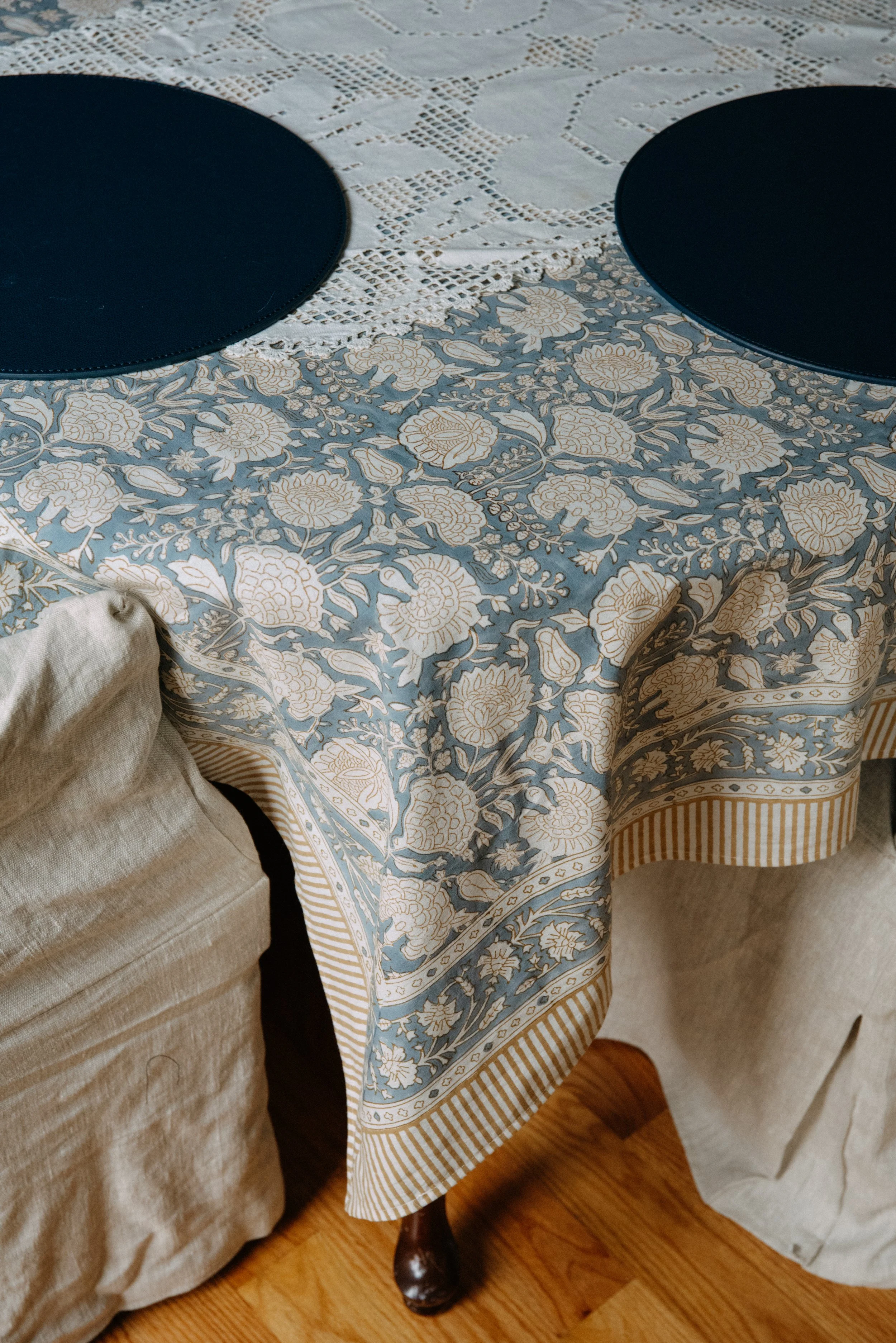 A dining table with a blue floral tablecloth, two black round placemats, a beige upholstered chair with a wooden leg, and part of a beige chair on a wooden floor.