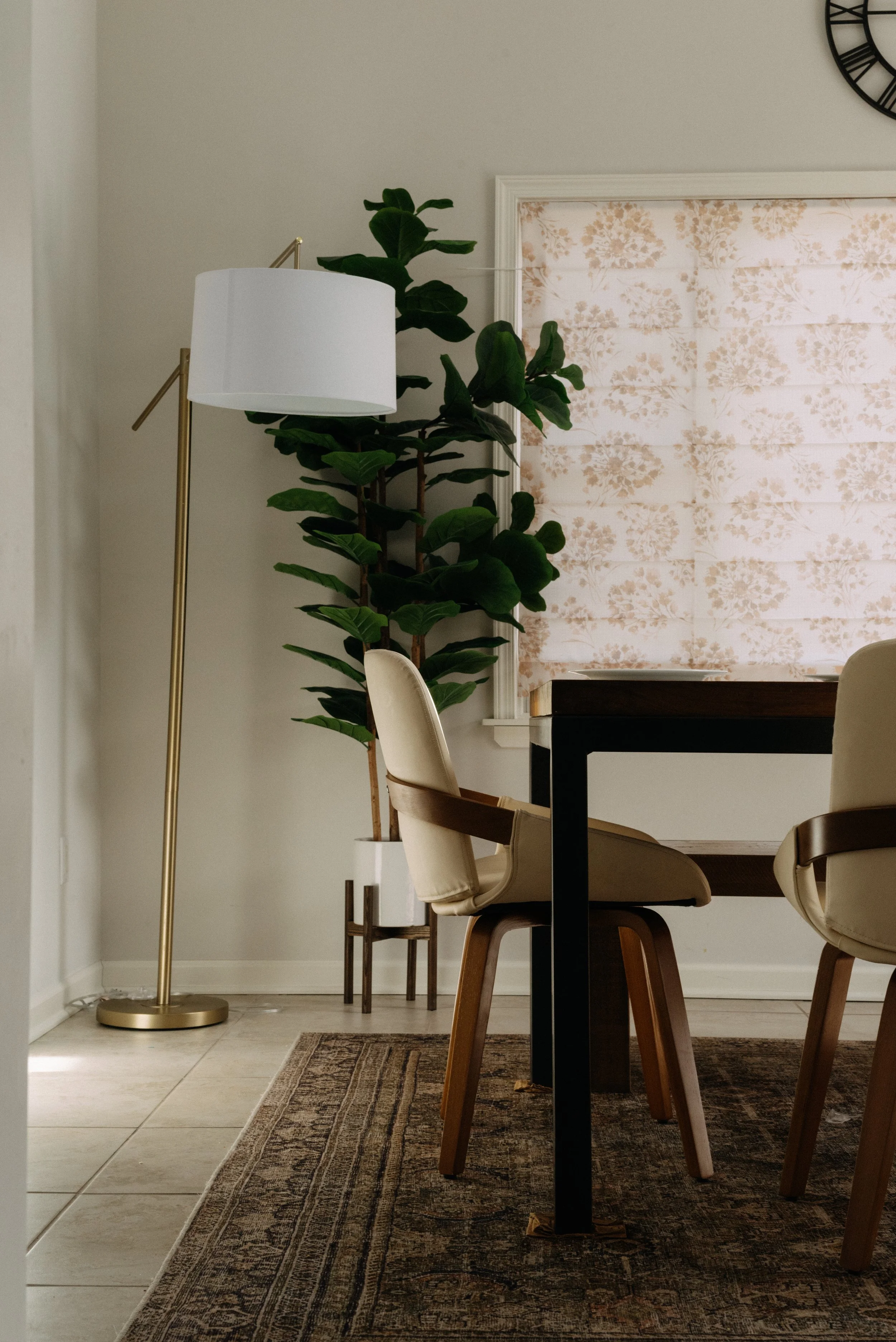 Dining room with beige upholstered chairs, a dark wooden table, a potted plant, a white and gold floor lamp, a window with a floral patterned shade, and a patterned rug.