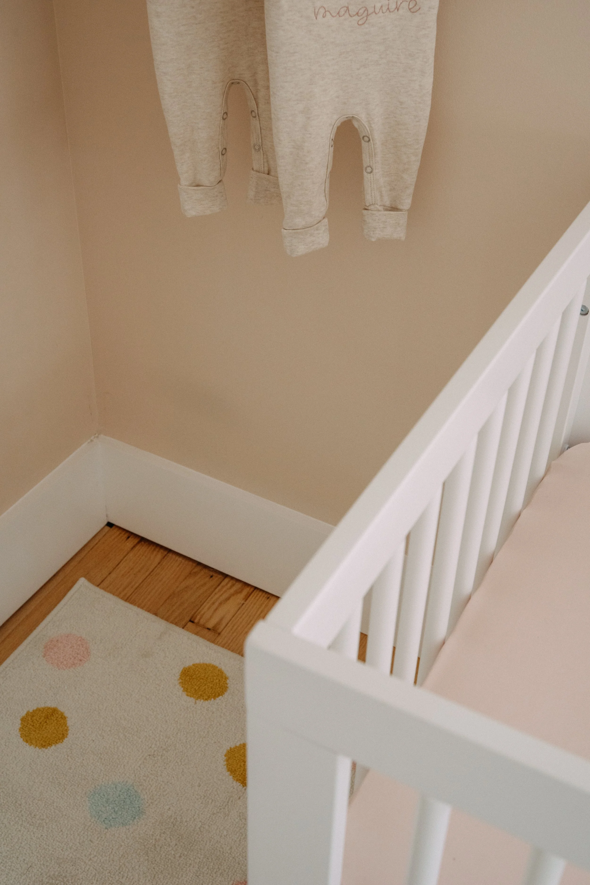 A corner of a nursery with a white crib, a beige wall, a soft gray baby onesie hanging on the wall, and a light-colored polka dot rug on a wooden floor.