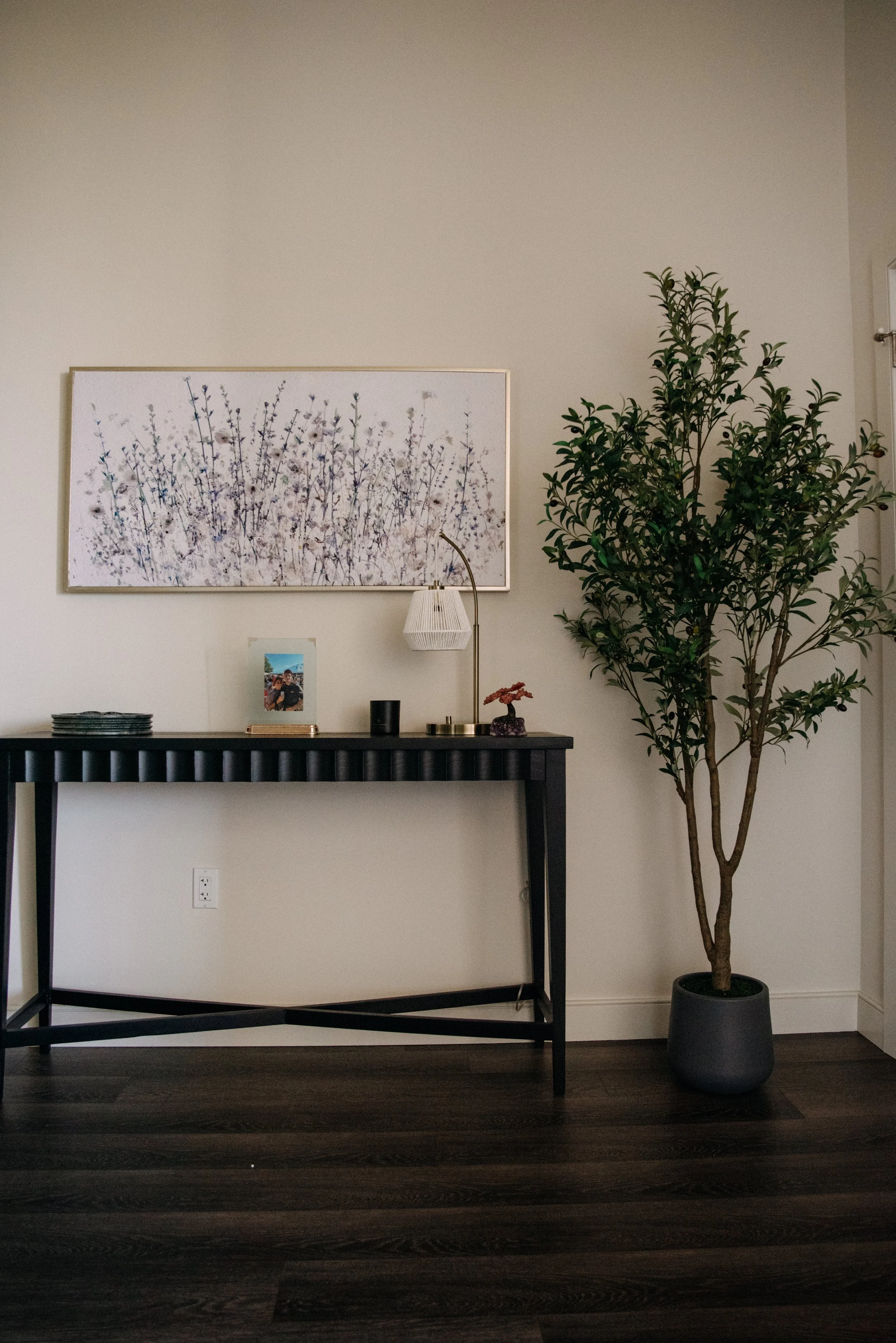 Interior of a room with a black console table, framed artwork of white and purple flowers, photo frame, black candle, white lamp, decorative pink flower, and large potted tree on dark hardwood floor.