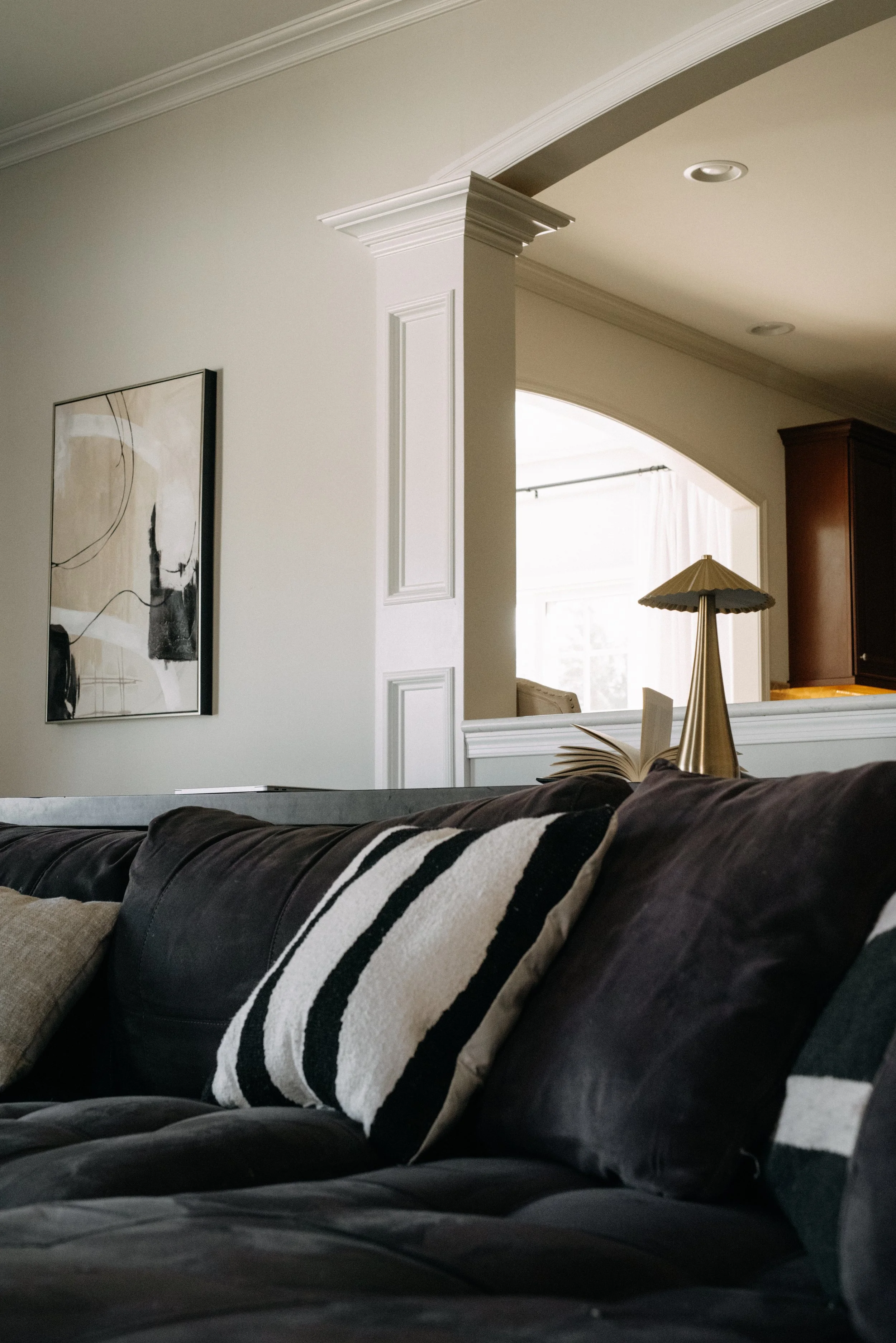 A living room with a dark leather sofa and decorative pillows, part of a white wall with artwork, and a view into a kitchen area with a lamp and open book in the background.
