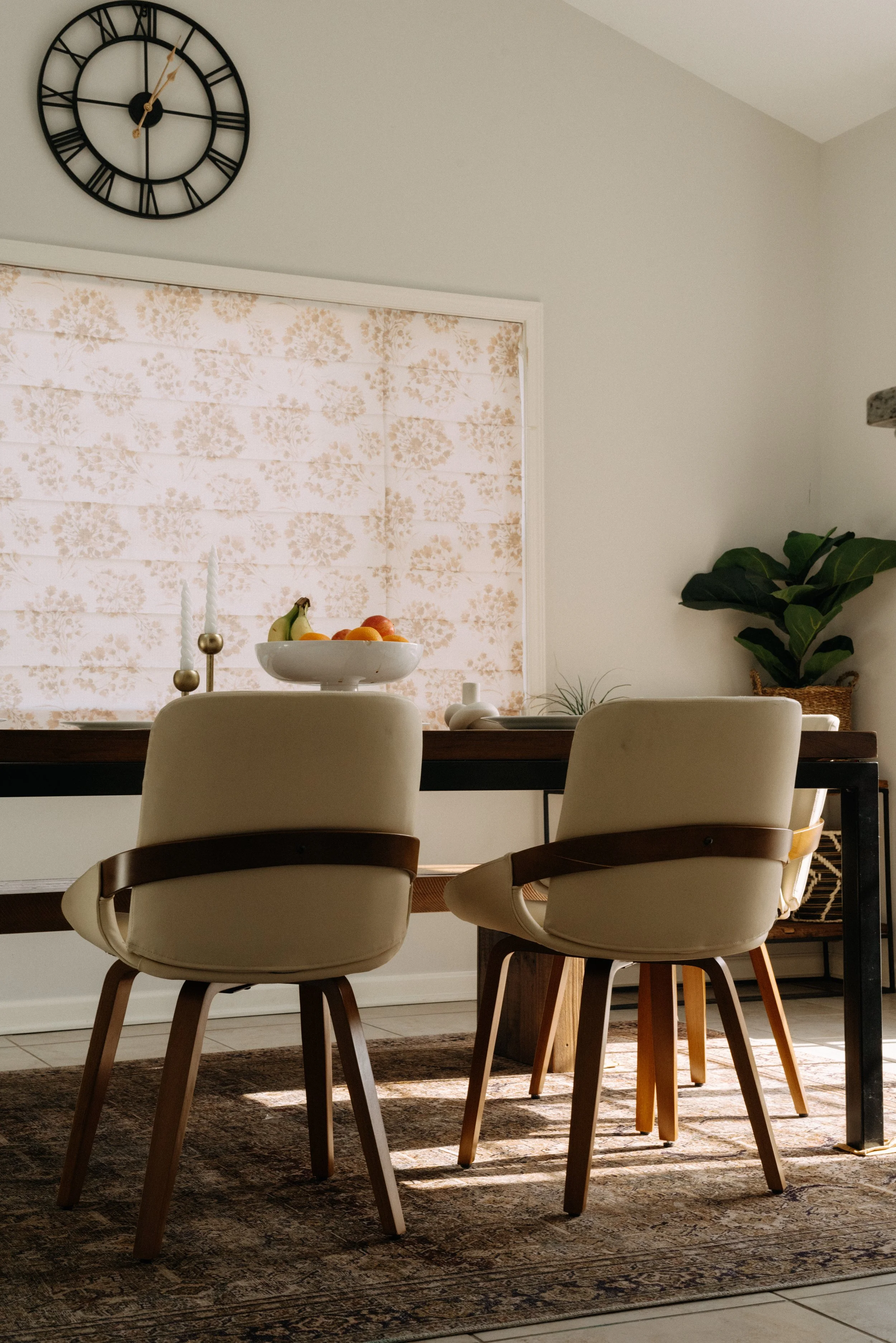 Dining room with beige chairs around a dark wood table, fruit bowl, two white candles with gold accents, potted plant, clock on wall, window with floral shade, and patterned rug.