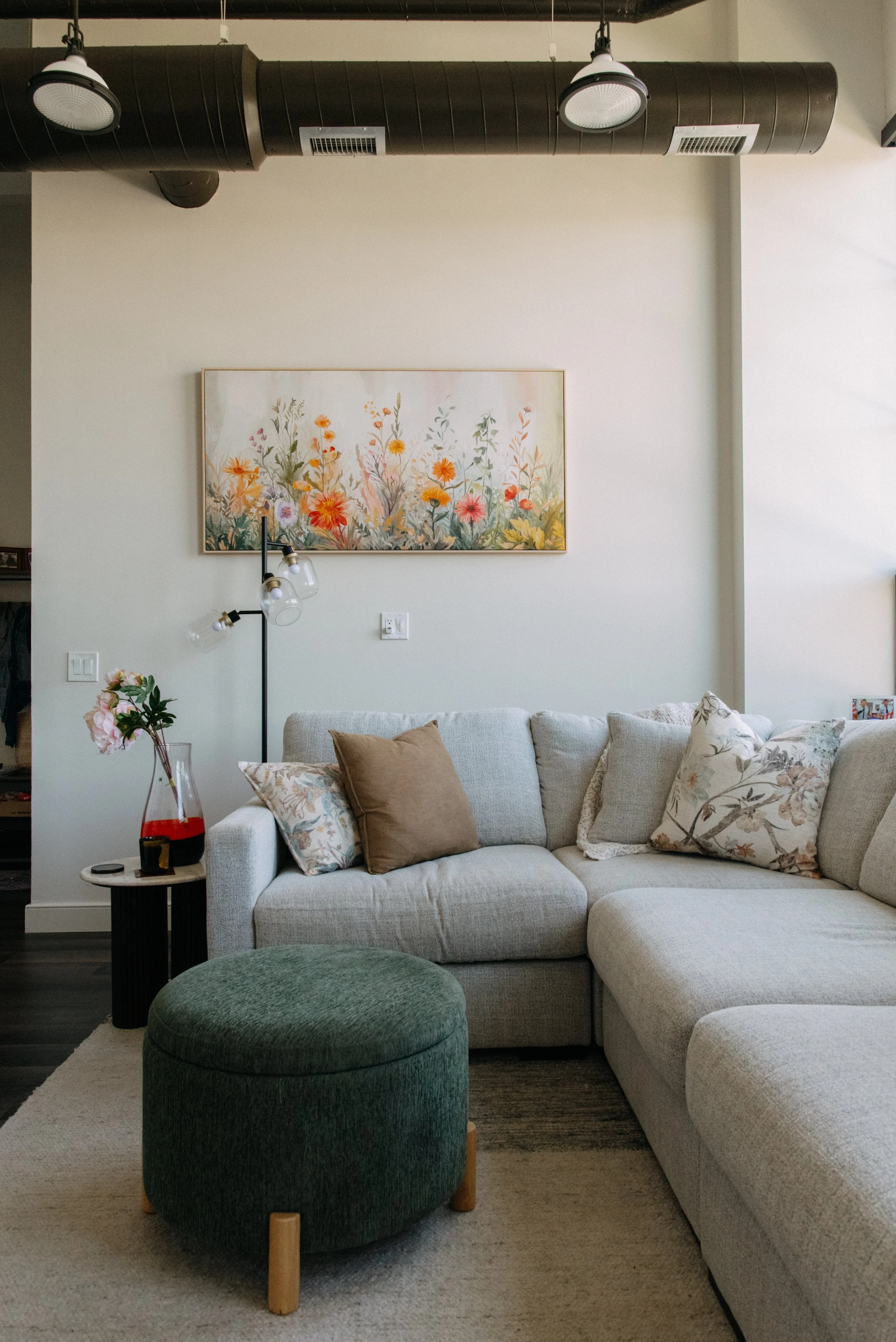 Living room with a beige sectional sofa, floral throw pillows, a round green ottoman, a side table with a flower vase, a tall floor lamp, a painting of flowers on the wall, a white wall, and an industrial black ceiling duct.