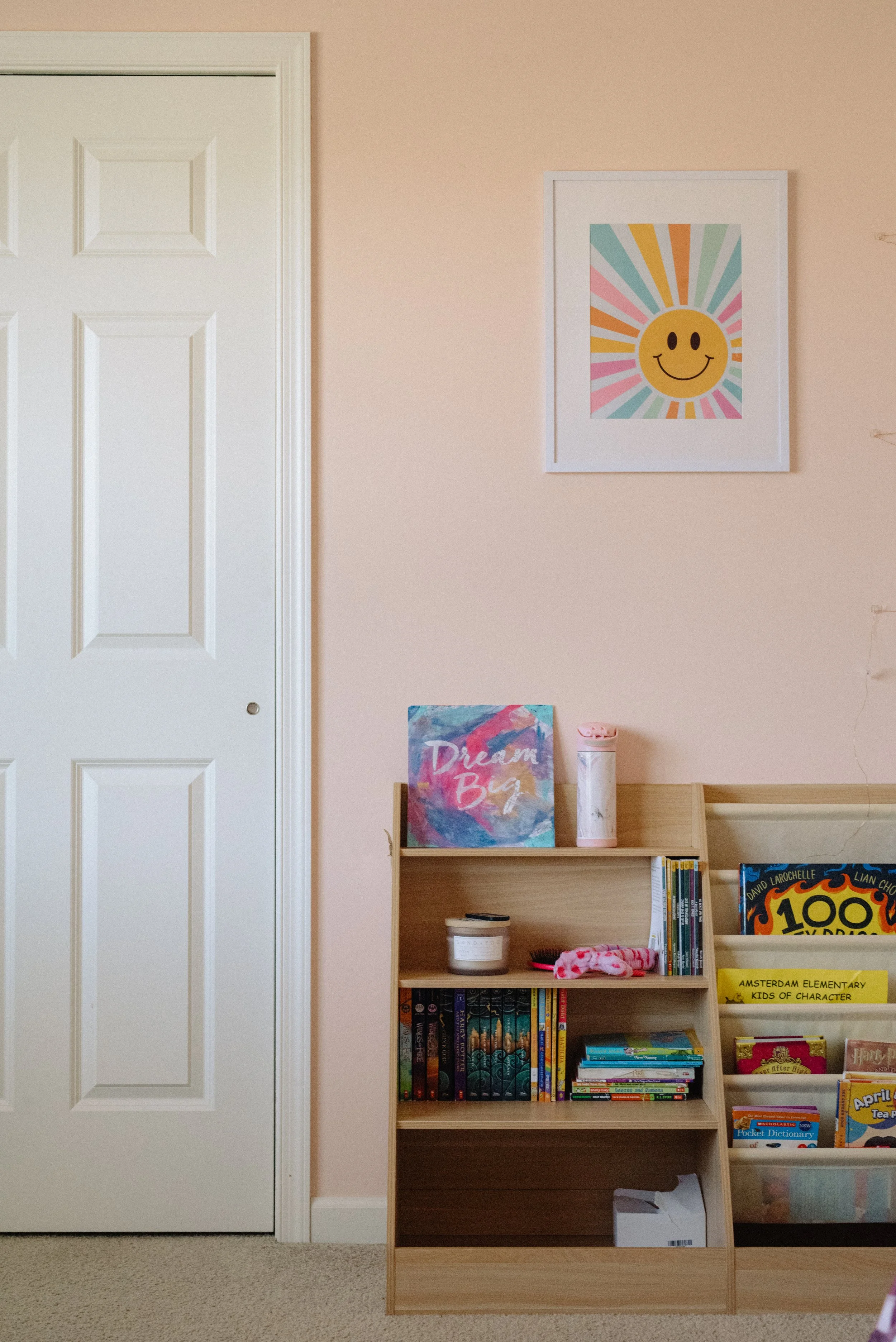 A bookshelf with children's books, a pink water bottle, and a colorful sign that says 'Dream Big,' positioned against a light pink wall with framed art of a smiling sun with rainbow rays.