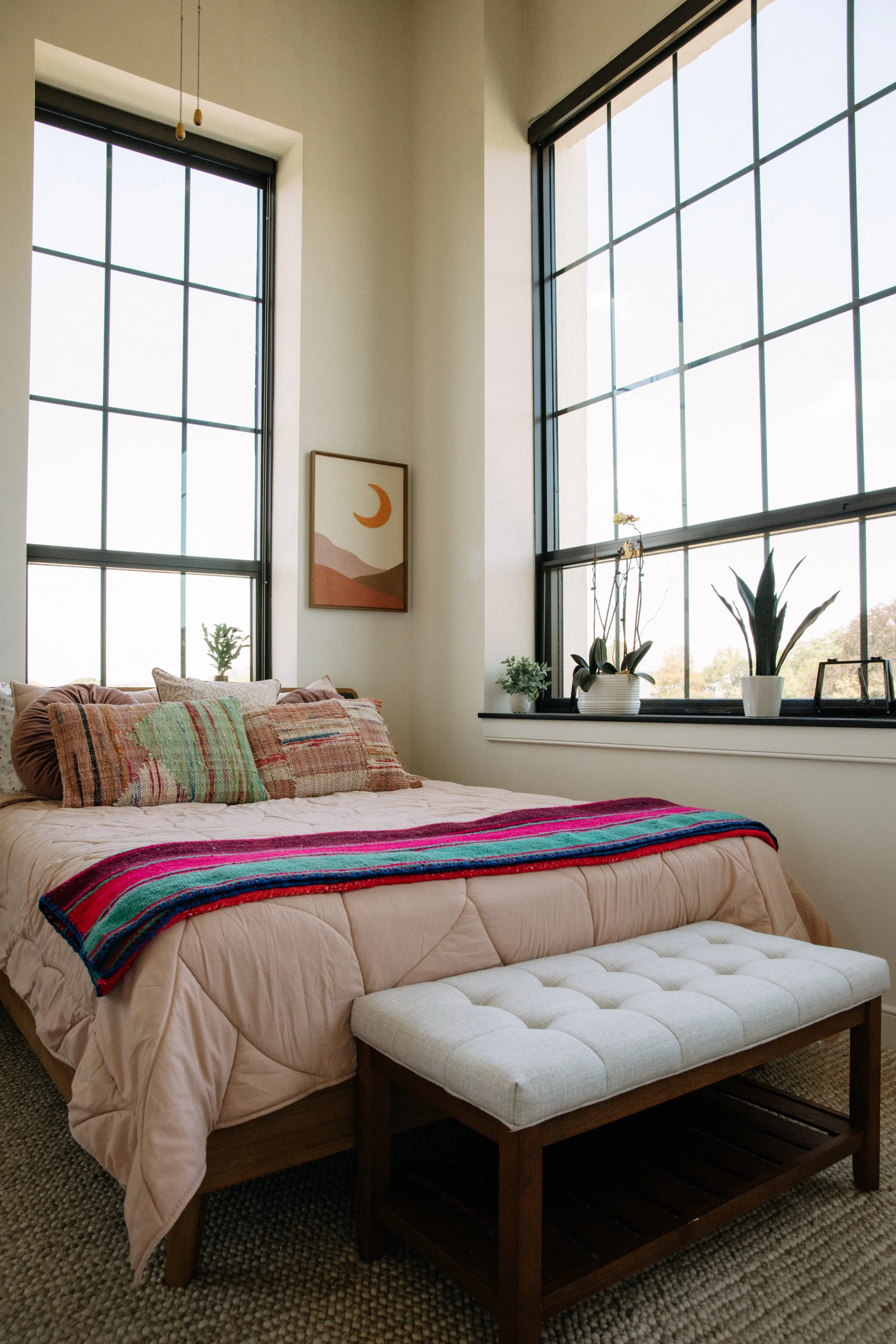 A cozy bedroom with a large window, decorated with plants, pillows, and a colorful blanket on the bed, a framed wall art featuring a moon, and a tufted bench in front of the bed.
