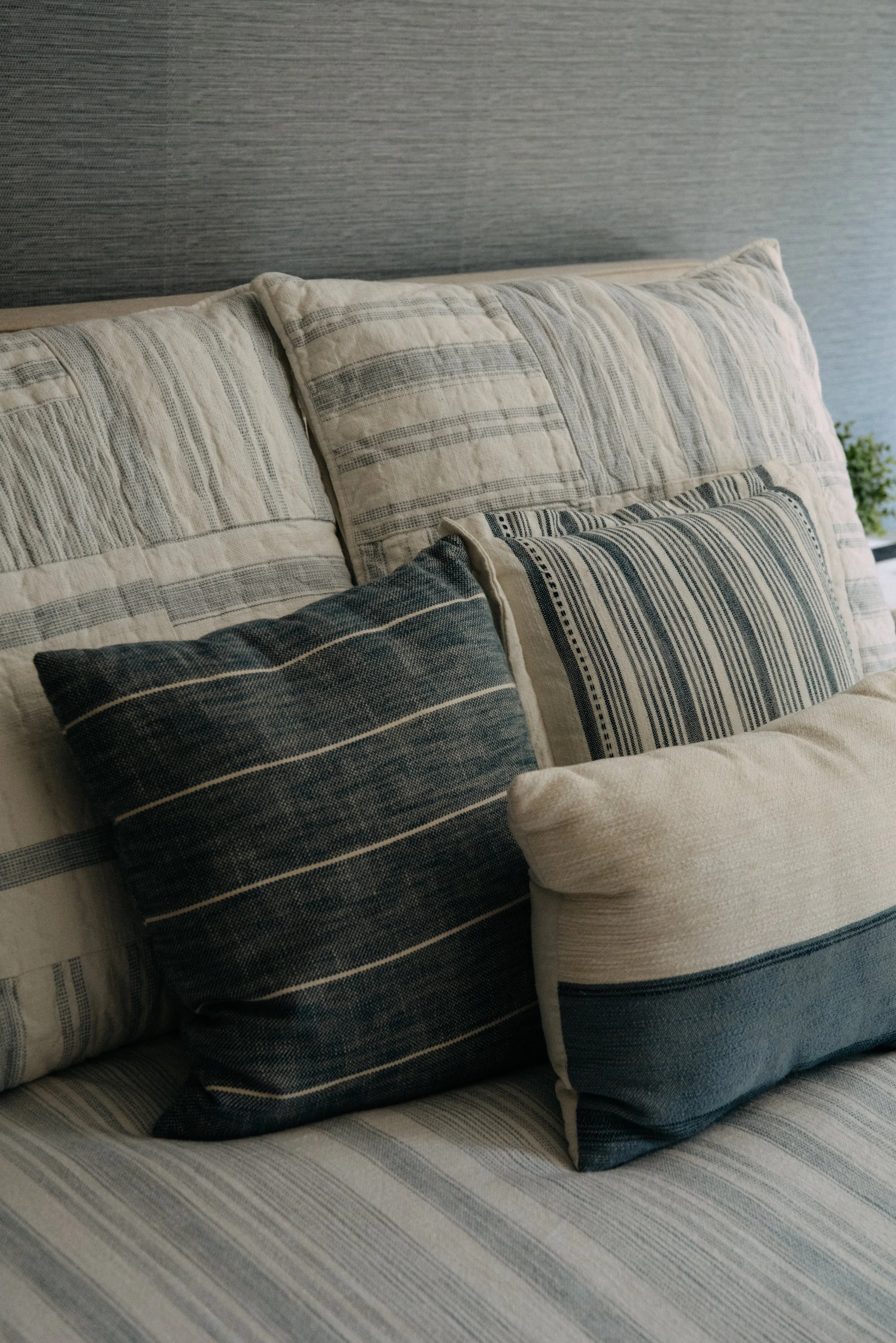 Close-up of a bed with multiple pillows featuring striped patterns in shades of beige, gray, and black, against a textured gray wall.