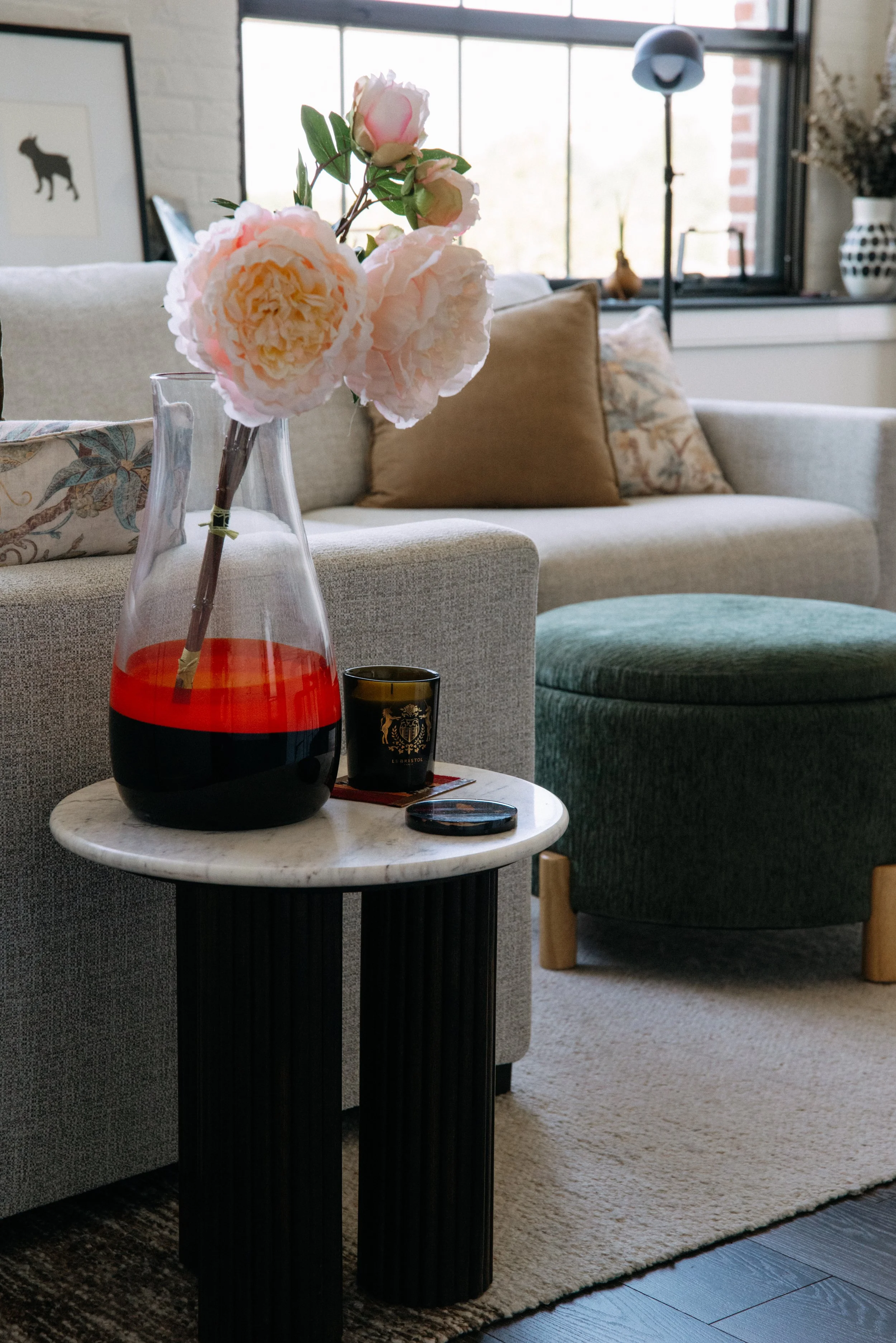 Living room with a vase of pink peonies on a white marble-topped table, a beige sofa with decorative pillows, and a green ottoman in front of a large window.