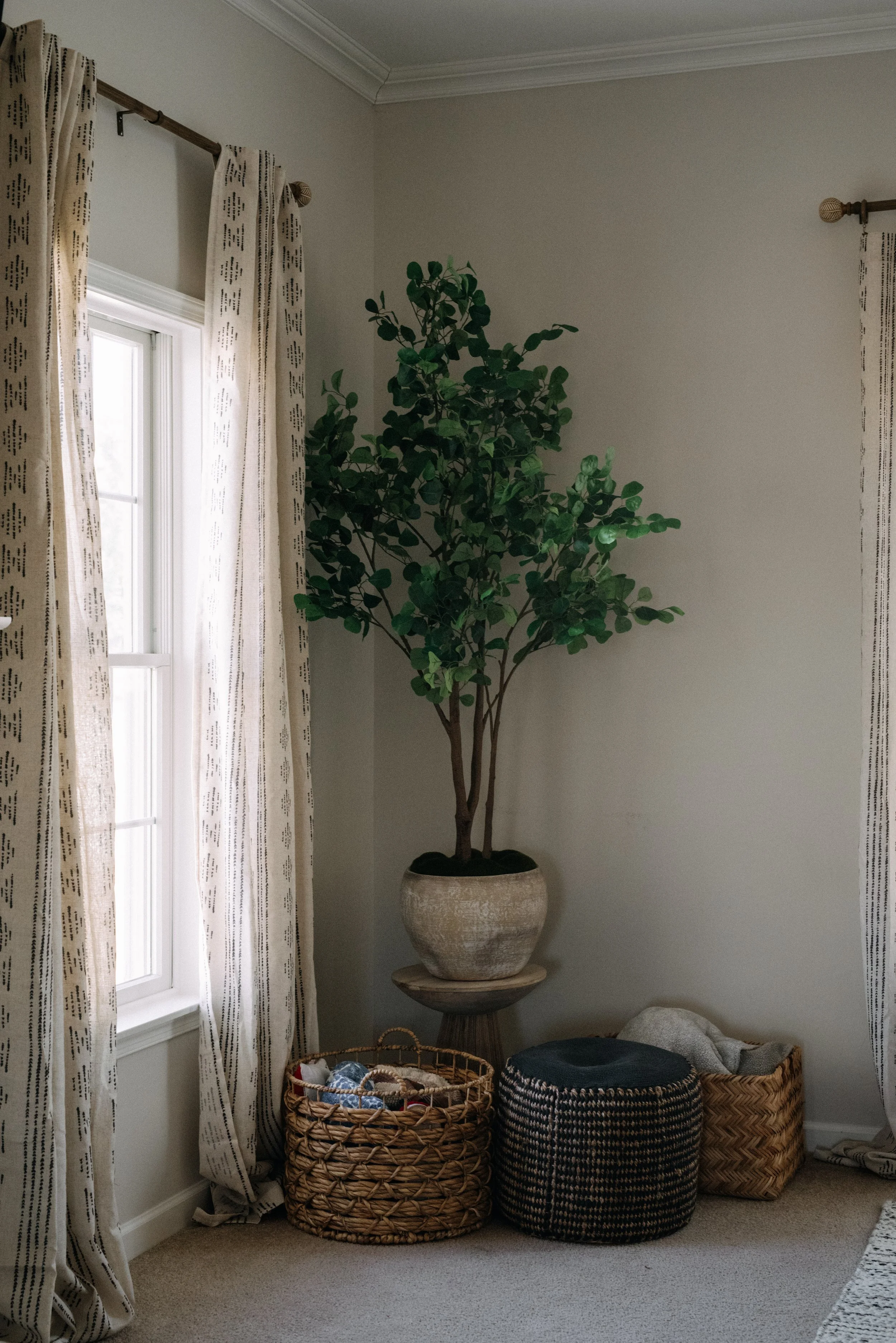 A tall potted green plant in a beige pot on a wooden stool next to a window with beige curtains. Rattan and fabric baskets are on the floor around the plant.