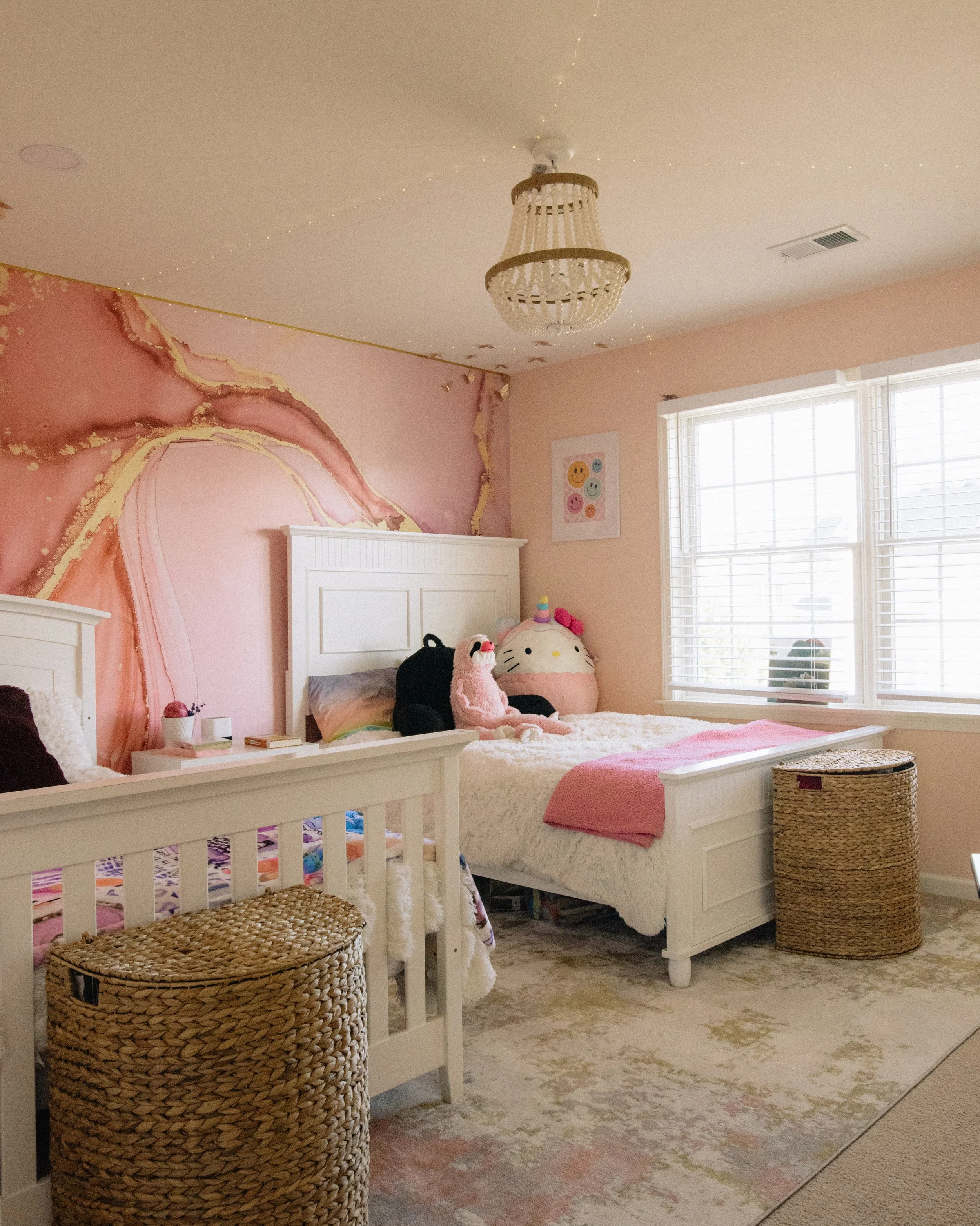 A bedroom with pink walls, white beds, plush toys including a unicorn and Hello Kitty, two woven baskets, a chandelier, and large windows with white blinds.