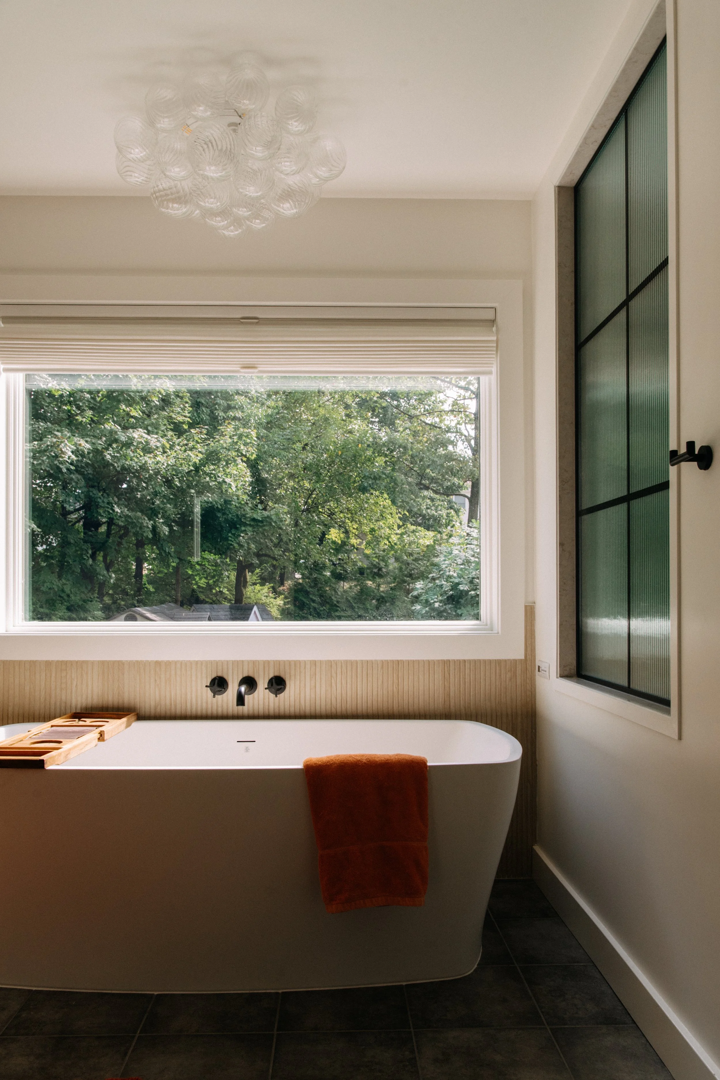 A modern bathroom featuring a freestanding bathtub with a wooden tray, orange towel, large window with greenery outside, and a stylish ceiling light.