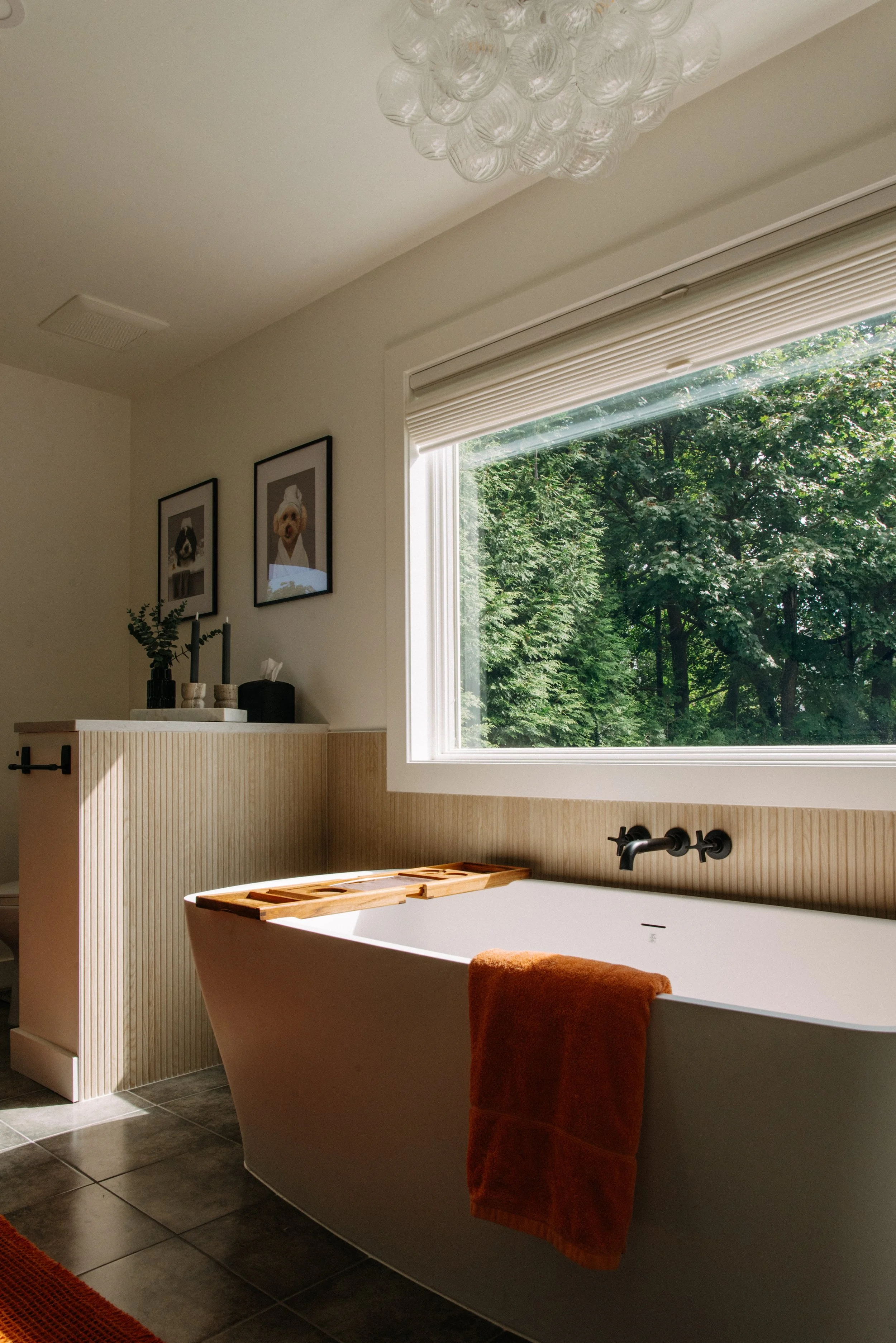 Modern bathroom with a white freestanding bathtub, a large window showing green trees outside, and framed pictures of dogs on the wall.