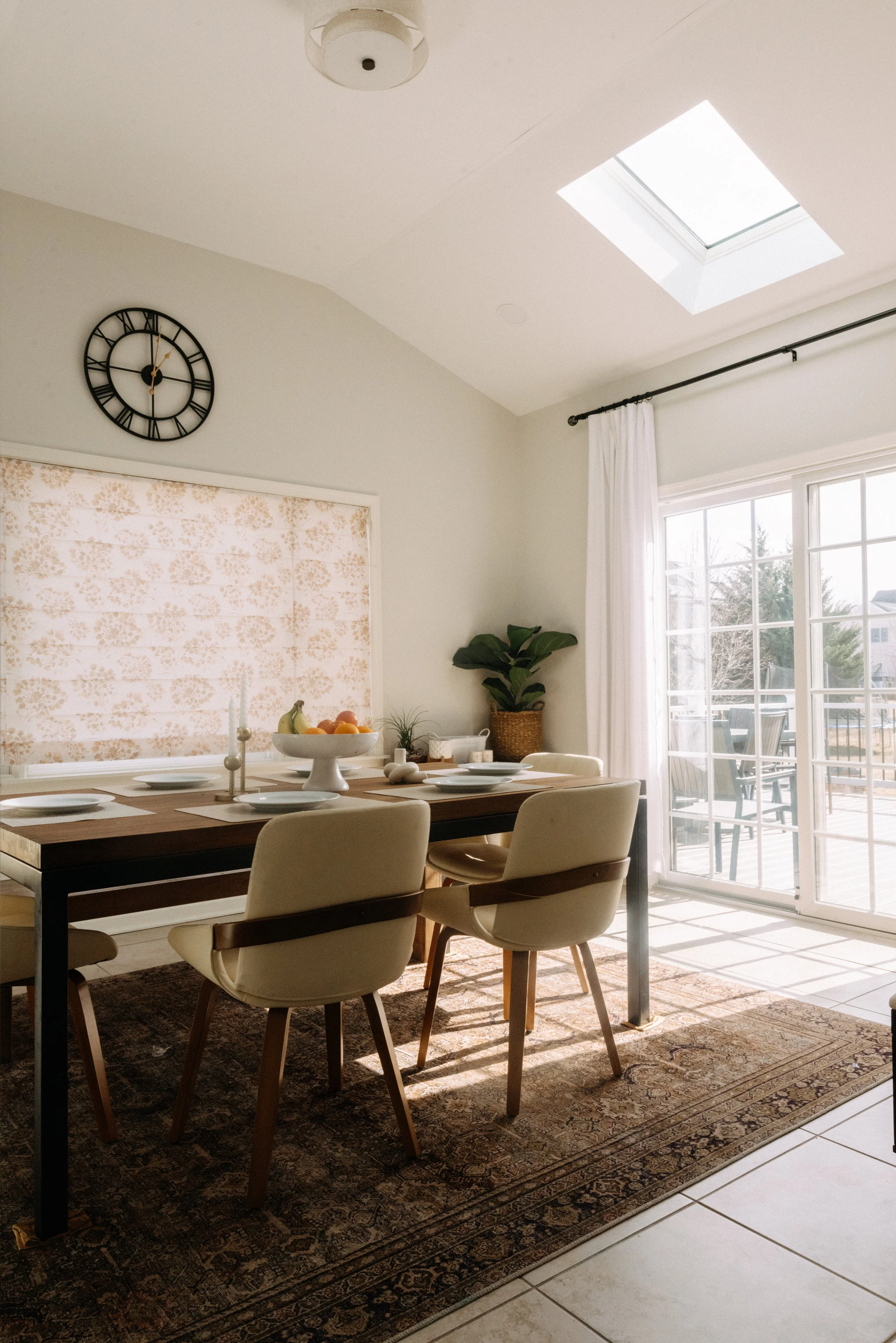 A dining room with a wooden table set with white plates, a bowl of fruit, and white candles, on a patterned rug. There are four cream-colored chairs, a large window with white curtains, a potted plant, a wall clock, and a skylight letting in natural