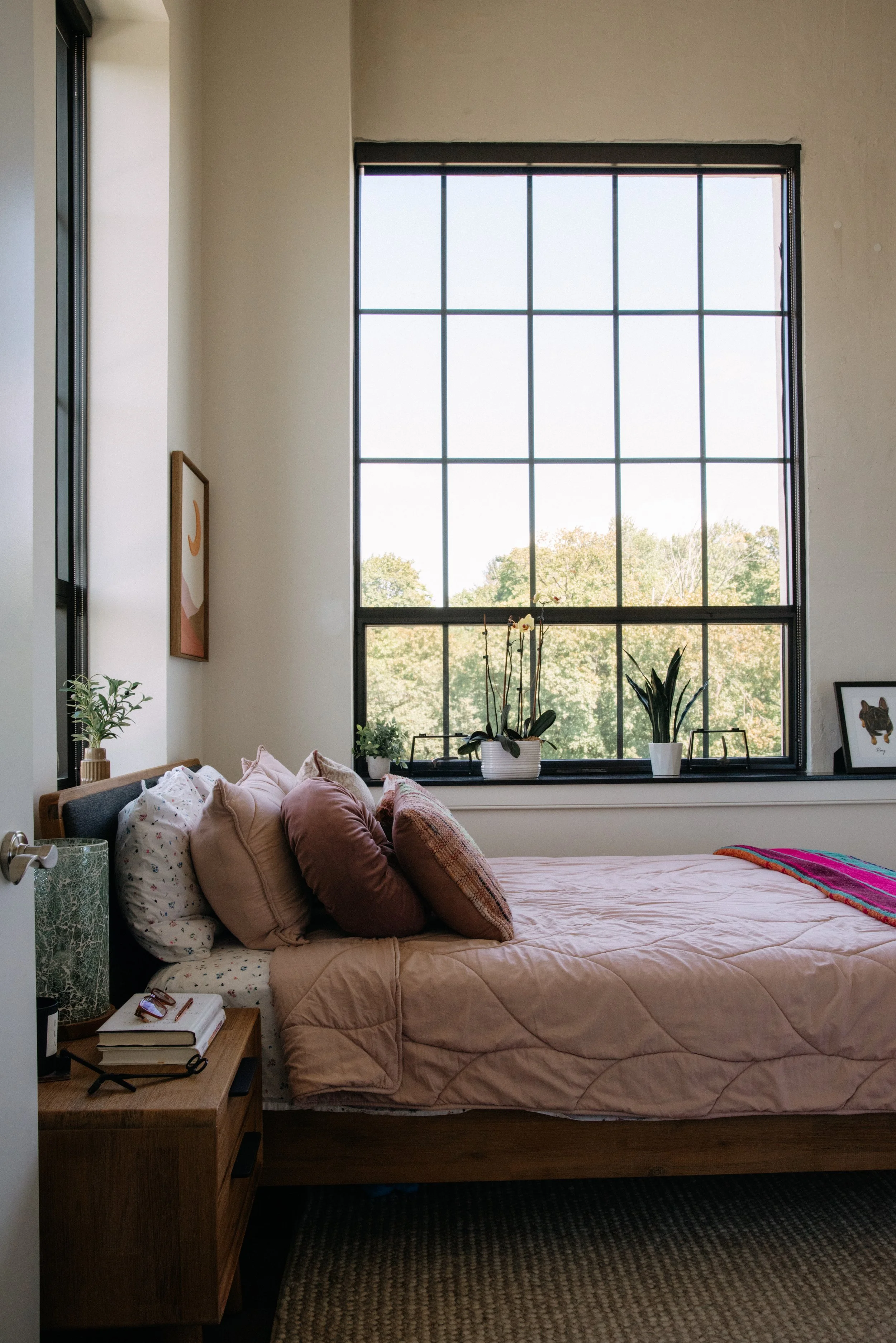 A bedroom with a large window showing trees outside, decorated with potted plants on the windowsill, a bed with a pink quilt and multiple pillows, a wooden nightstand with books and glasses, and a framed art piece on the wall.