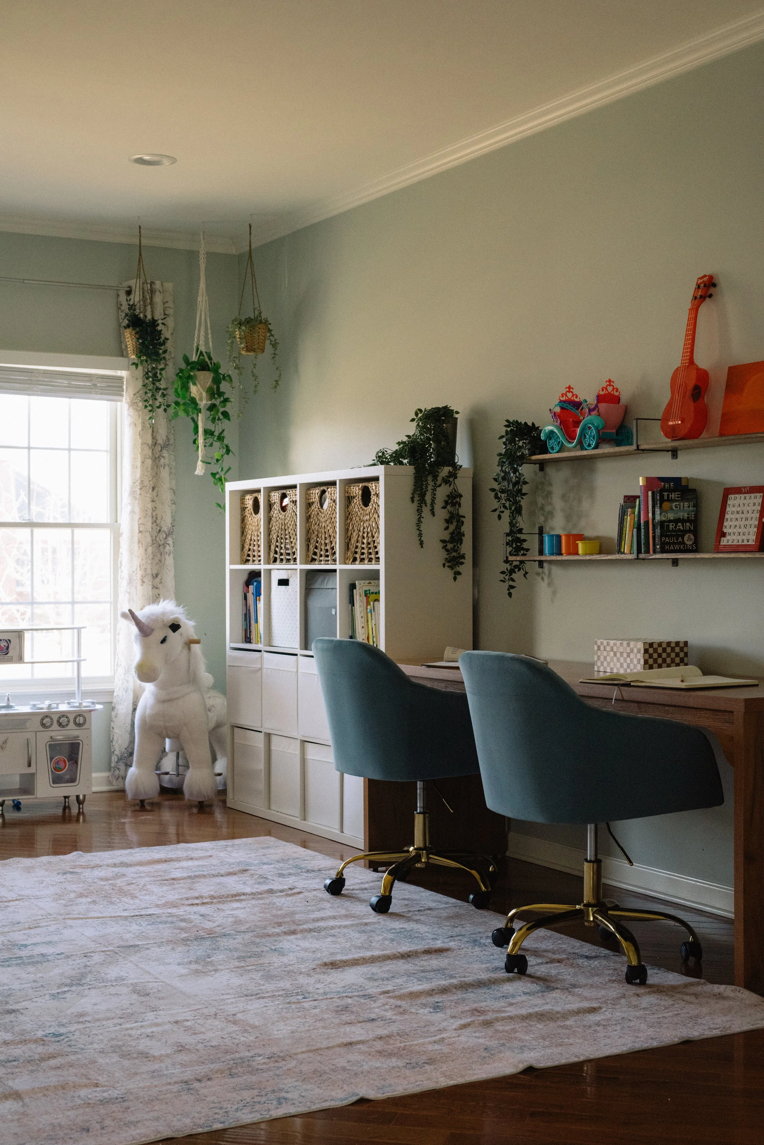 A home office or study space with a desk, two teal office chairs, and a white storage unit filled with books and baskets. There are hanging plants and a plush unicorn chair near a window, along with colorful toys and books on wall shelves.