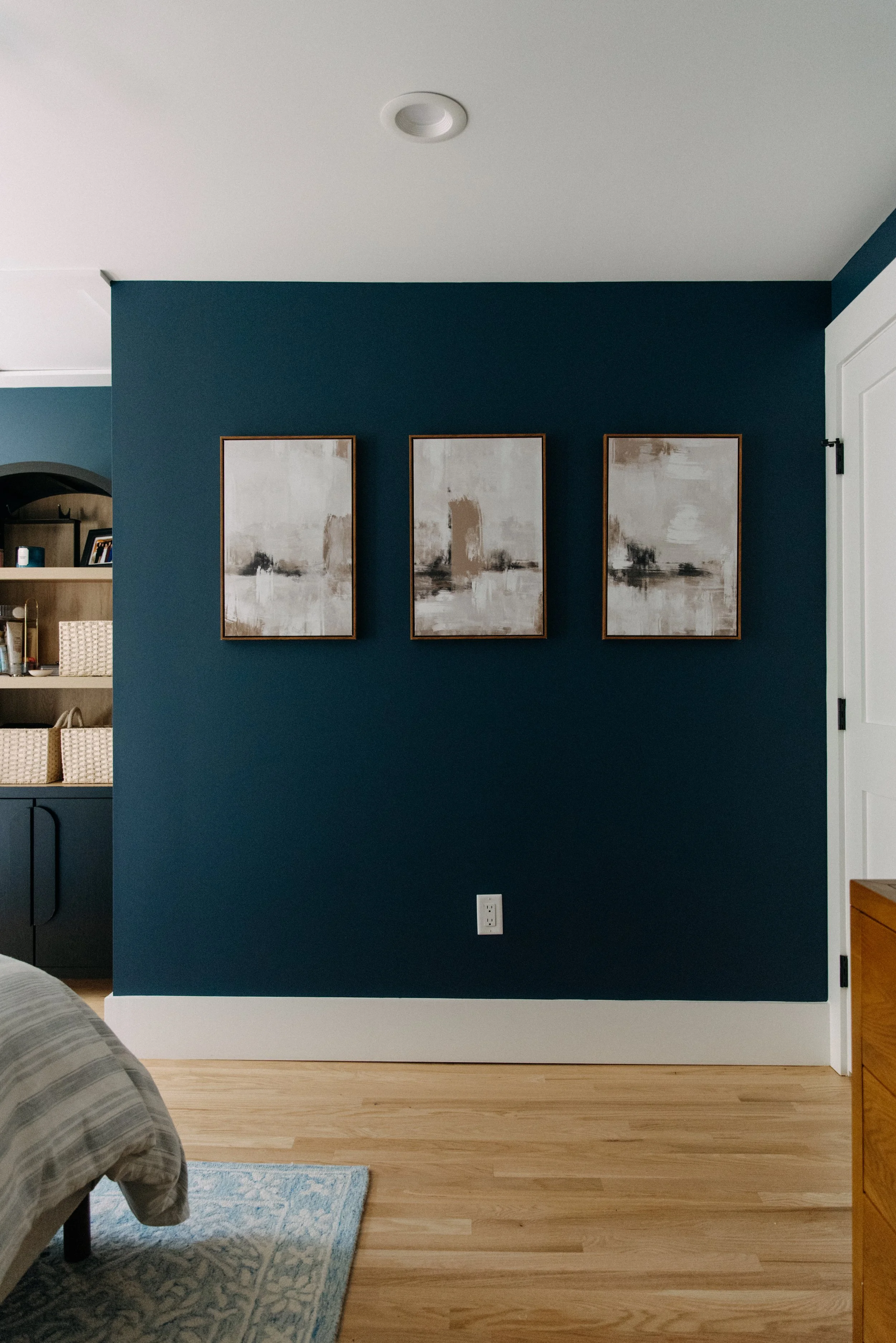 Interior view of a bedroom with a dark blue accent wall decorated with three abstract black and white paintings; part of a bed, a rug, and a wooden dresser are visible.