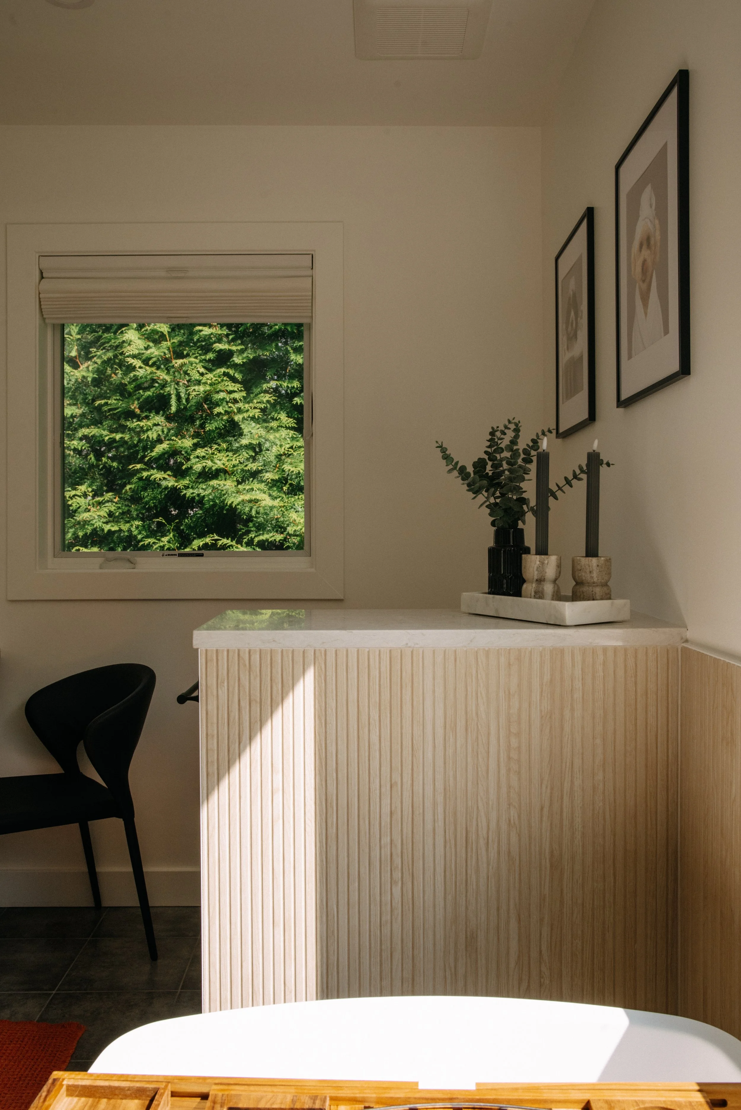 A cozy indoor space featuring a small window with lush green trees outside, a black chair, a wooden cabinet with a white marble top, two black candles in beige holders, a black vase with greenery, and framed artwork of dogs hanging on the wall.