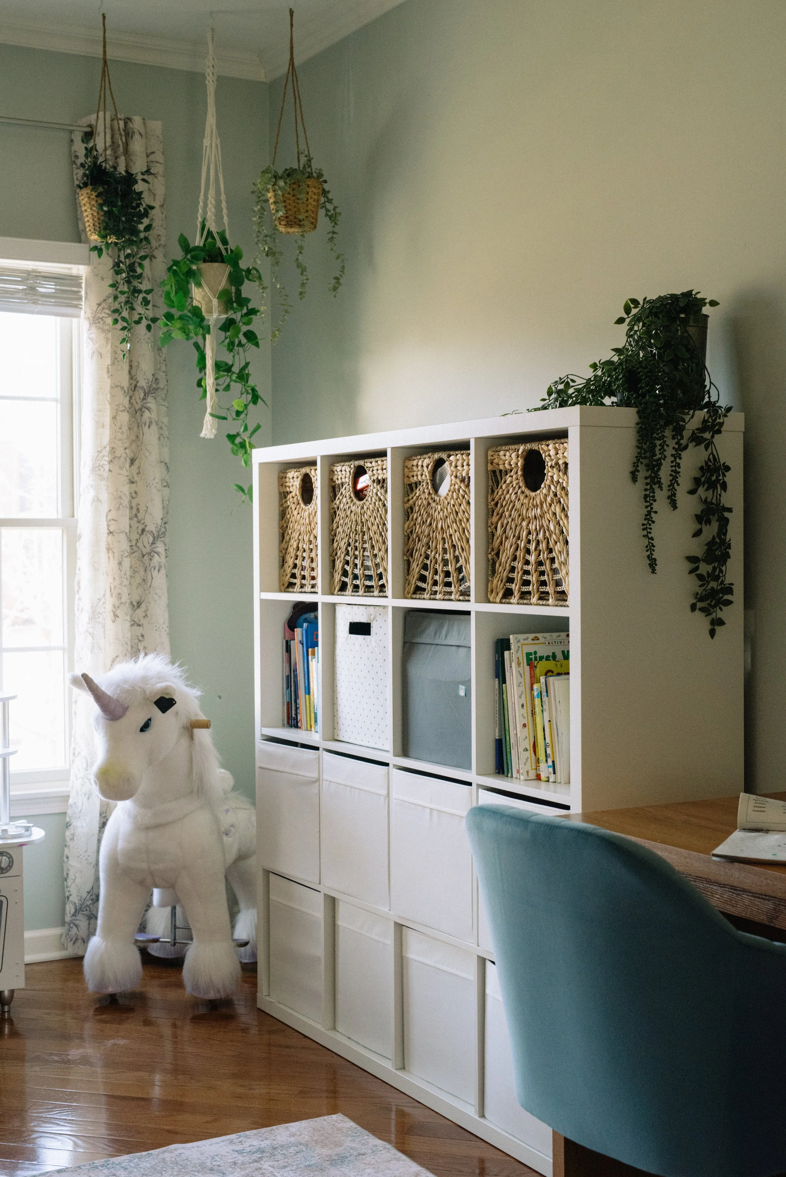 A white storage unit with woven baskets and books in a child's room, a large plush unicorn toy, hanging plants, and a window with curtains.